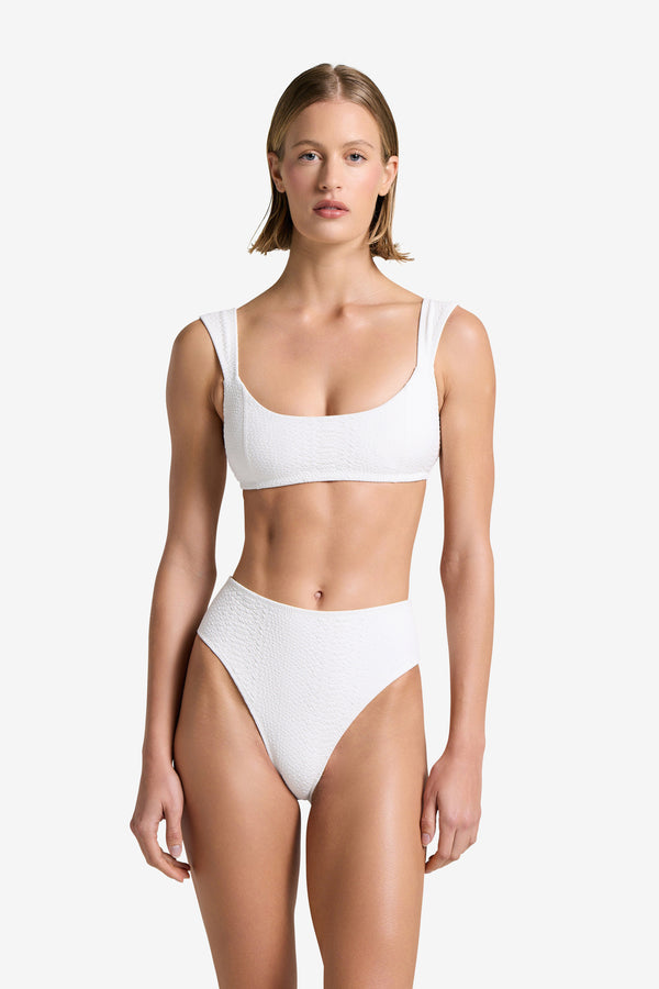 A person with shoulder-length hair stands against a plain background, wearing the Gigi C Bikinis Dawson Top. This white textured bikini is in snakeskin-embossed fabric with wide straps, removable bust pads, high-waisted high-cut bottoms, and is fully lined.