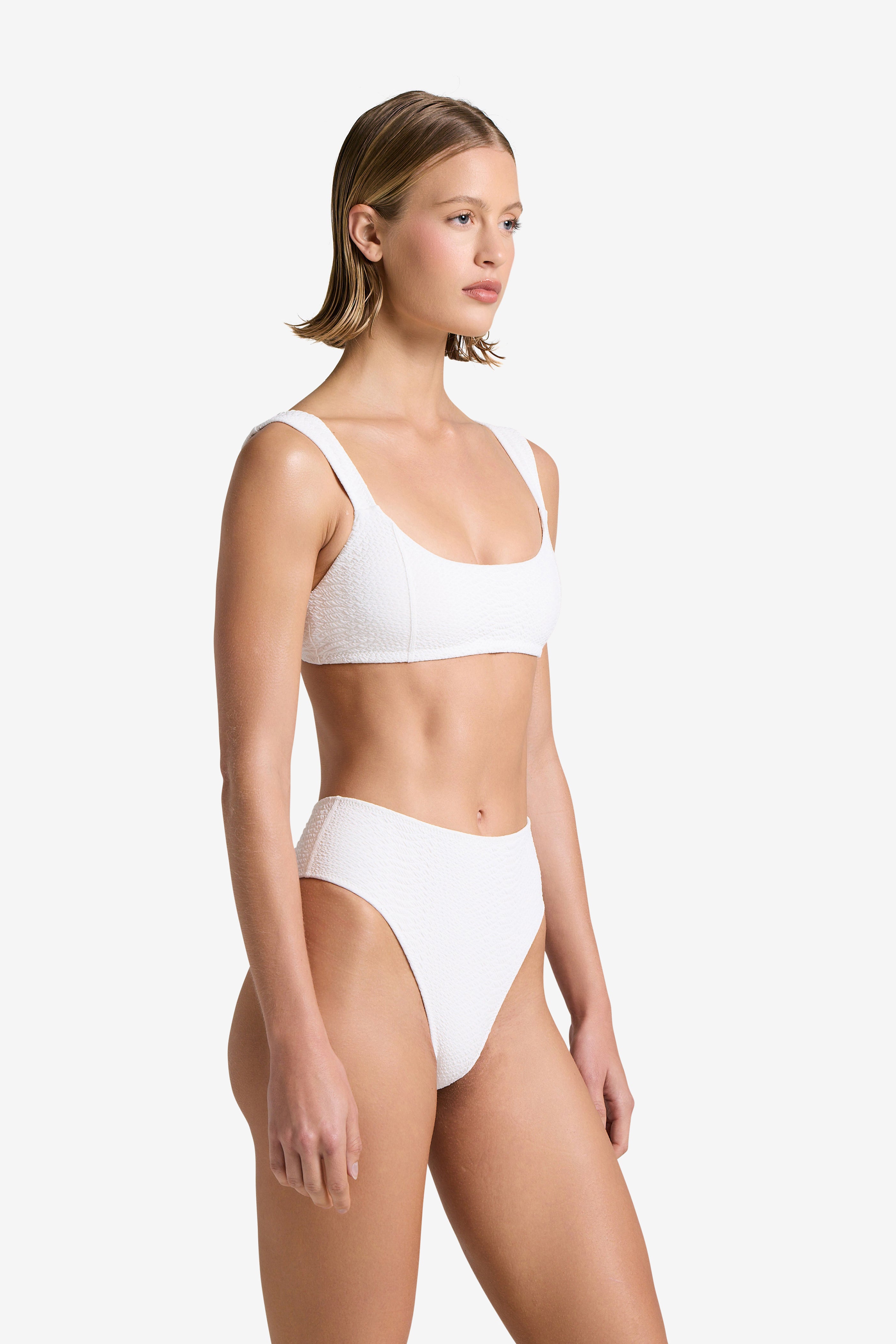 A person models Gigi C Bikinis Dawson Top, a white textured faux snake bikini with high-waisted bottoms and a scoop neck top, against a plain white background. They have short, straight hair and the bikini is elegantly accented with a subtle gold swan clasp.