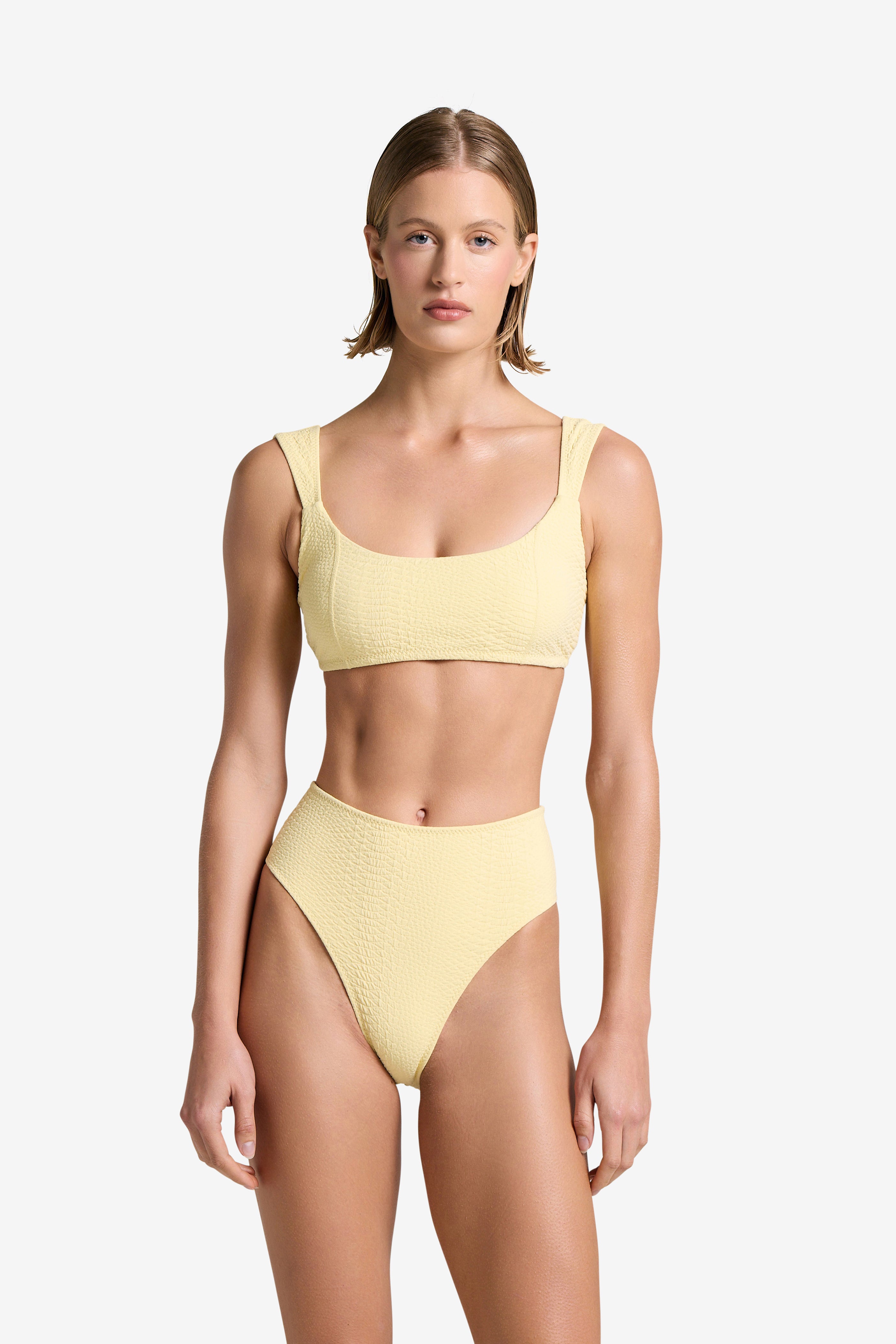 Wearing Gigi C Bikinis Dawson Top, a person faces forward in a yellow textured bikini with snakeskin embossed fabric, featuring a square neckline and high-waisted bottoms. Removable bust pads are included. The scene is set against a plain white background.
