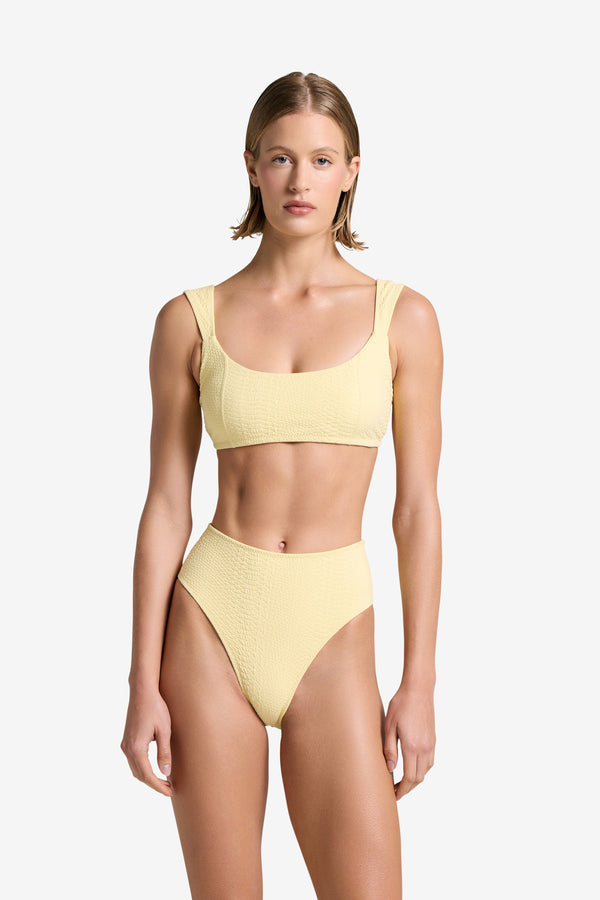 Wearing Gigi C Bikinis Dawson Top, a person faces forward in a yellow textured bikini with snakeskin embossed fabric, featuring a square neckline and high-waisted bottoms. Removable bust pads are included. The scene is set against a plain white background.