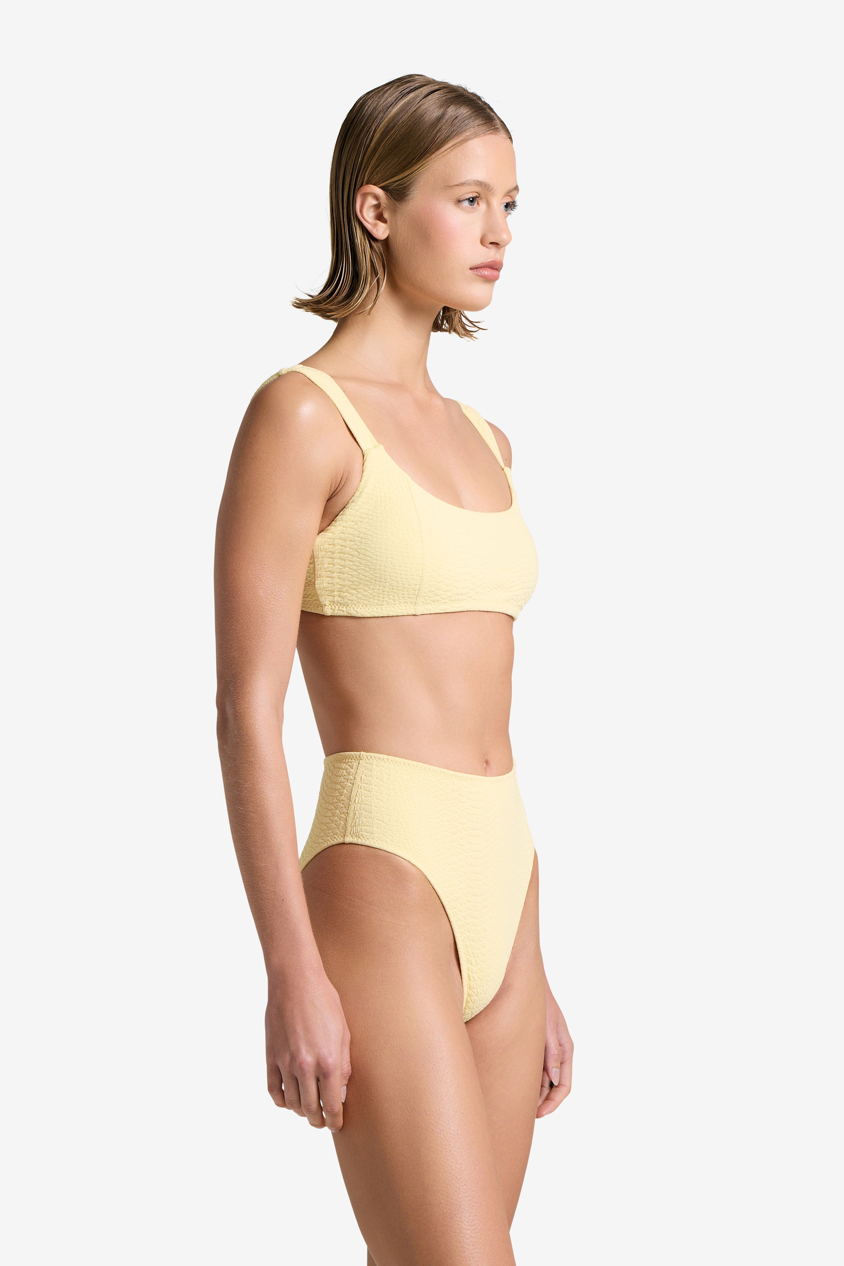 A woman with short hair models the Dawson Top by Gigi C Bikinis, a textured pale yellow bikini made from snakeskin-embossed fabric. She stands in side profile against a plain white background.