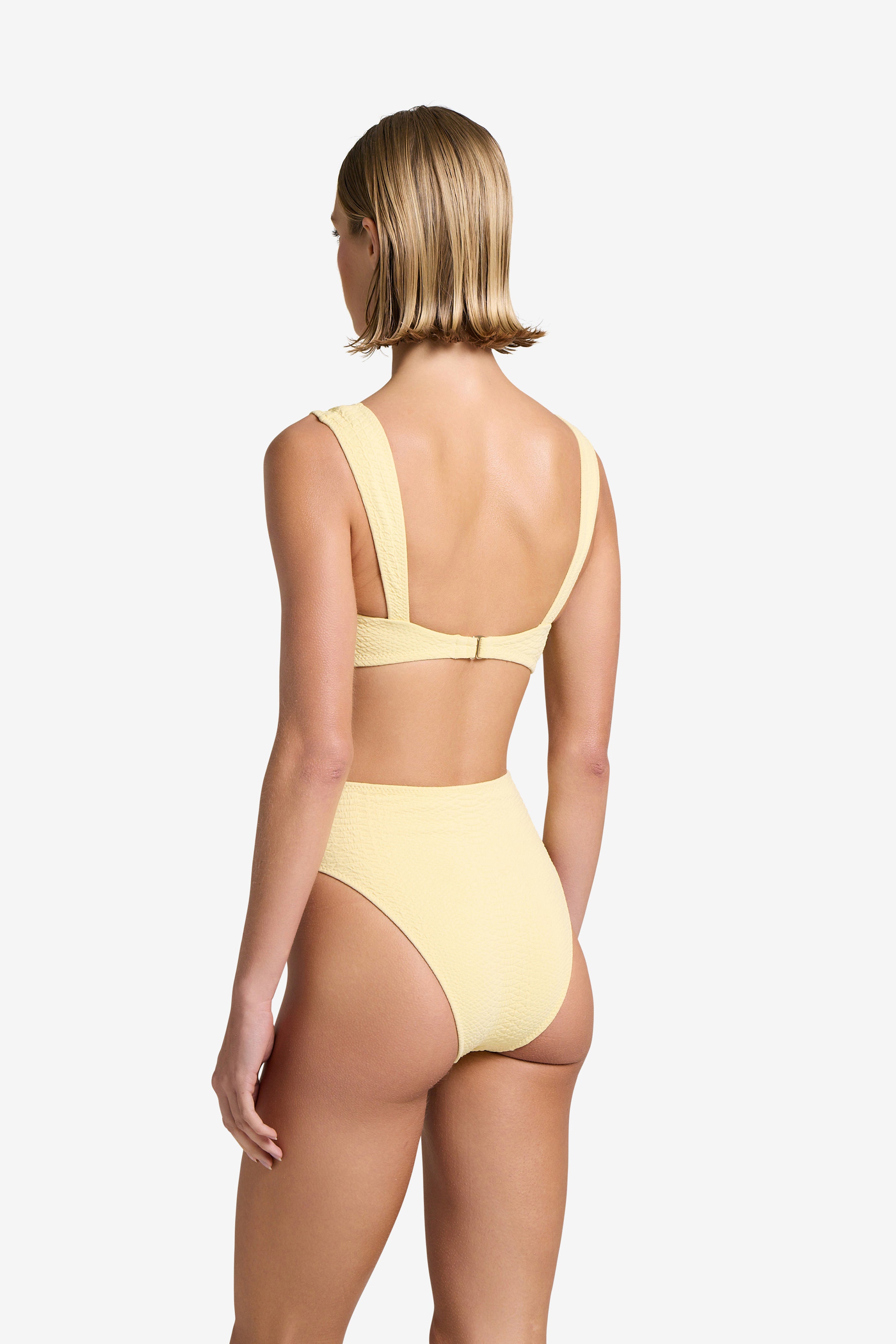 A person with short blonde hair is seen from the back wearing a textured yellow snakeskin-embossed bikini by Gigi C Bikinis. The Dawson Top has wide shoulder straps and a clasp, paired with high-waisted bottoms against a plain white background.