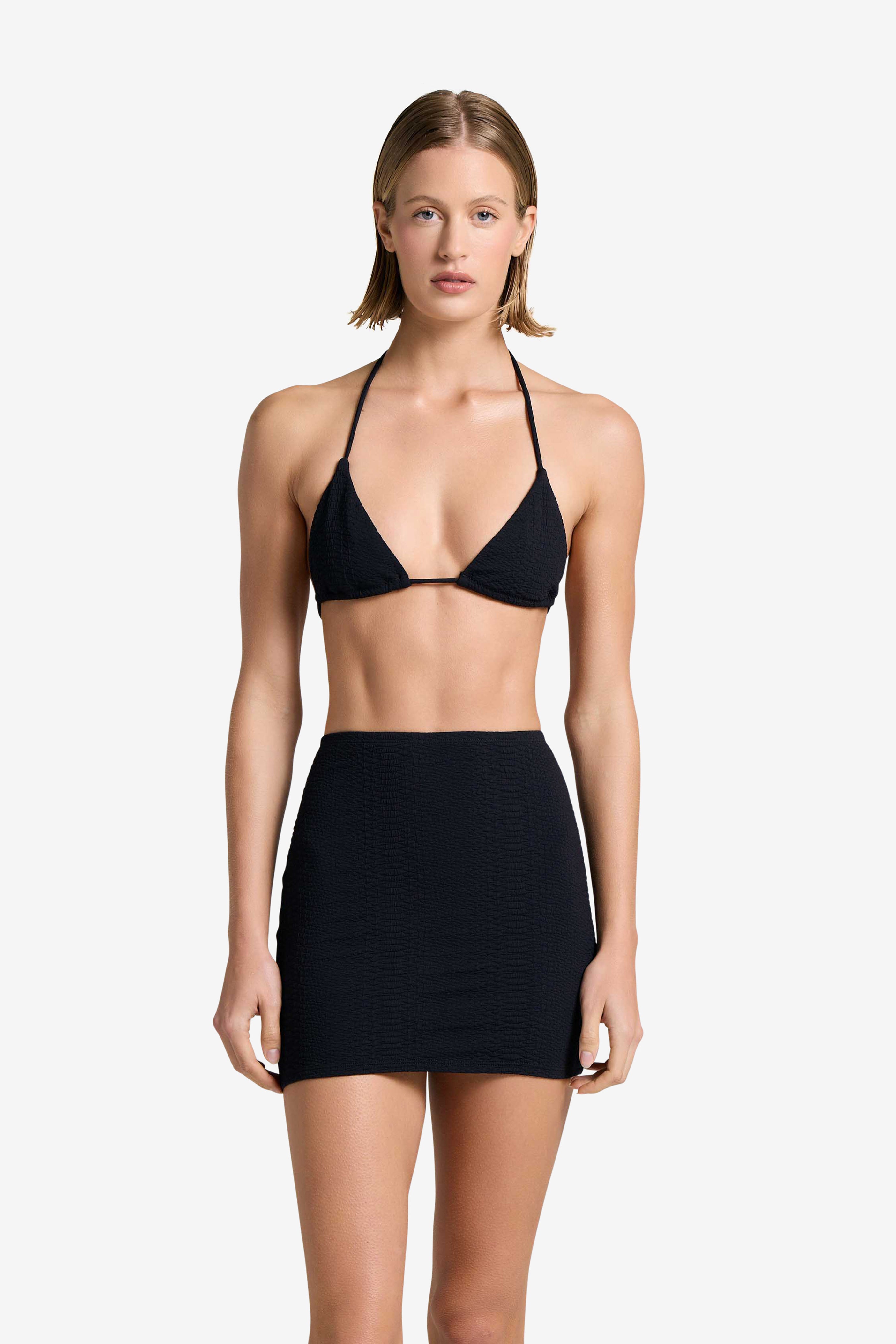A person with short hair is wearing a black bikini top and the sleek, body-shaping Joelle Skirt from Gigi C Bikinis. They stand against a plain white background, looking forward with a neutral expression.