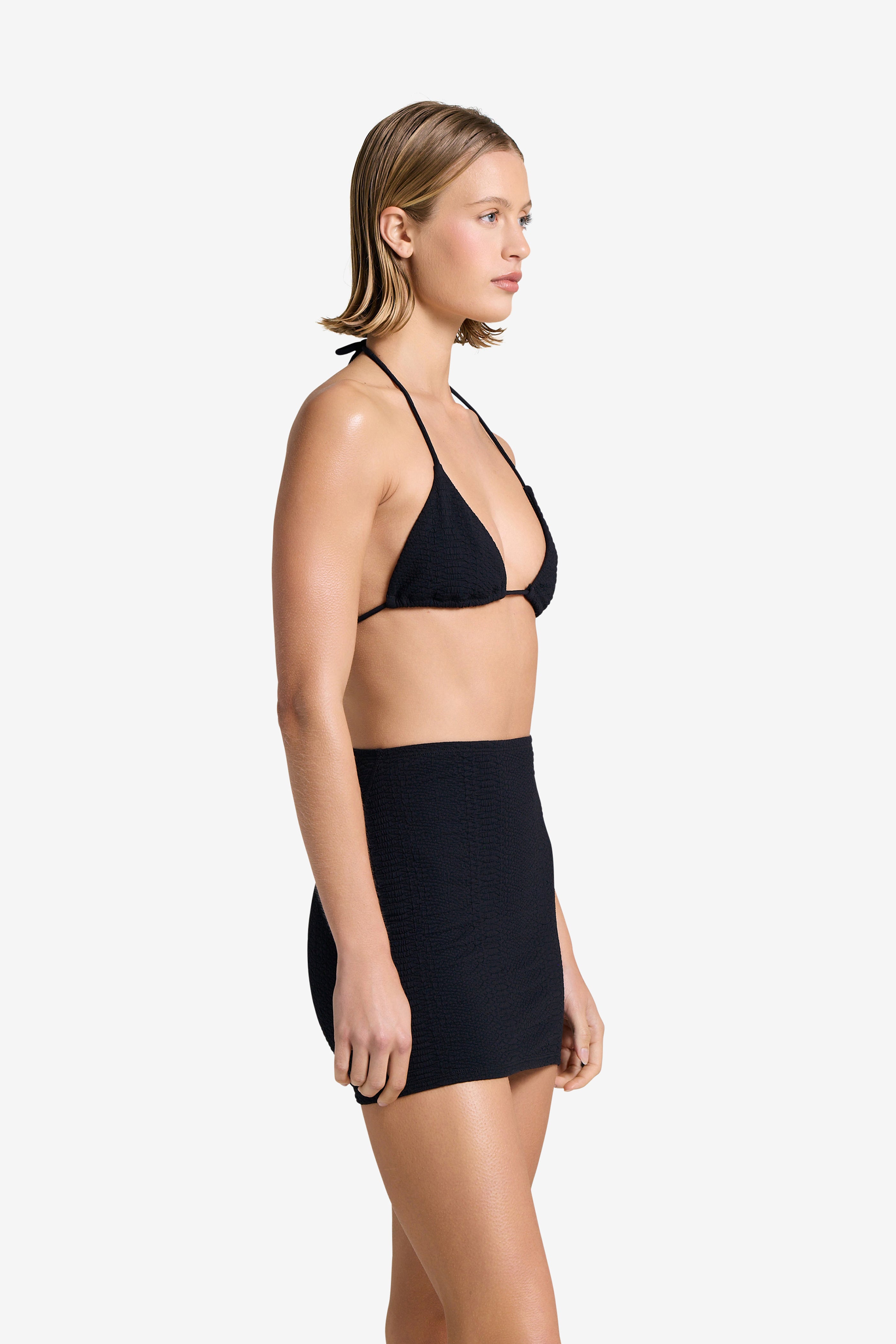 A person with short blonde hair is in side profile against a plain white background, wearing a black bikini paired with the sleek Joelle Skirt by Gigi C Bikinis.
