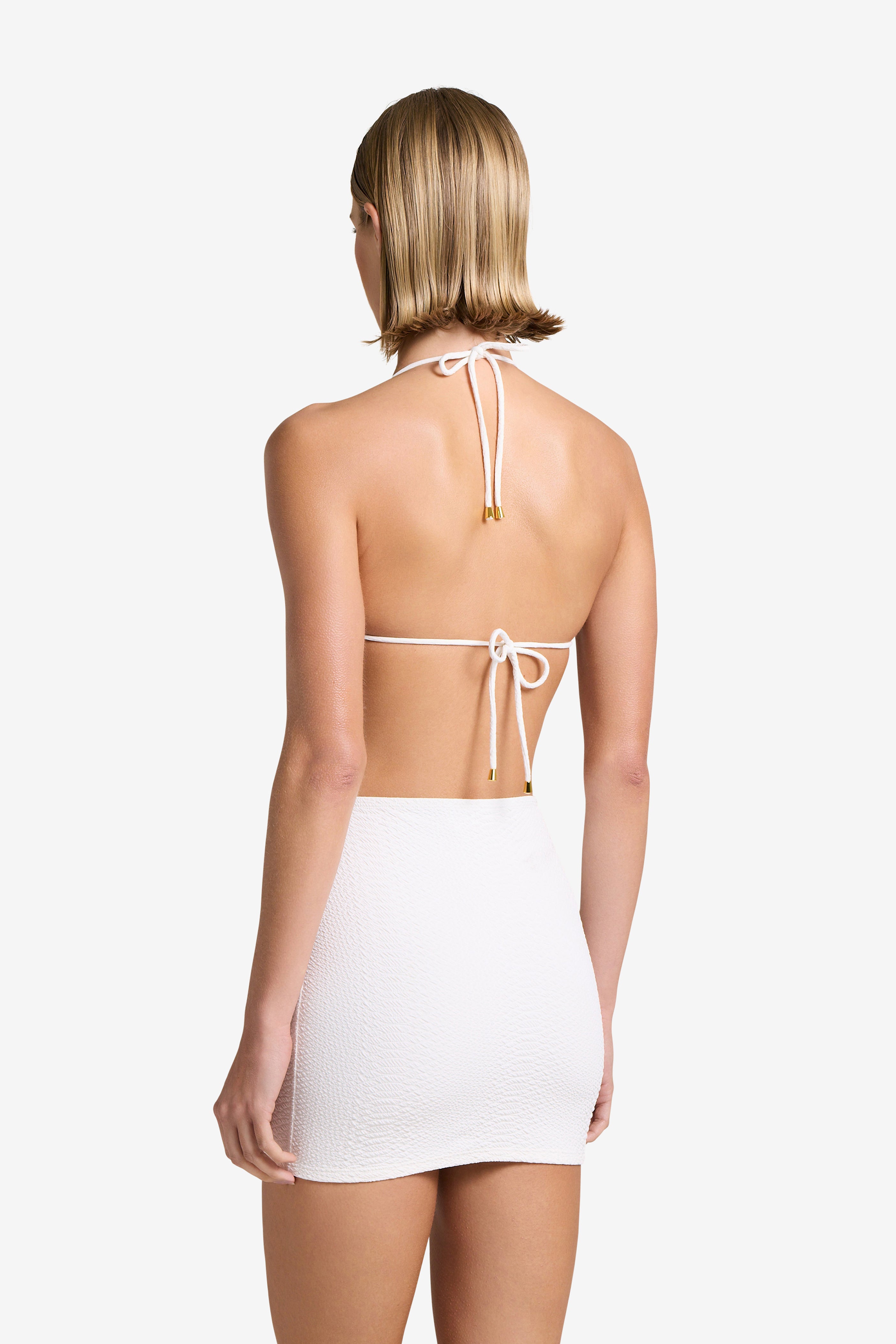 A person with short blonde hair faces away, wearing the Joni Top and a matching skirt by Gigi C Bikinis. The white halter tops thin straps tie elegantly at the neck and back, accented by gold hardware. The plain white background complements this chic summer ensemble perfectly.