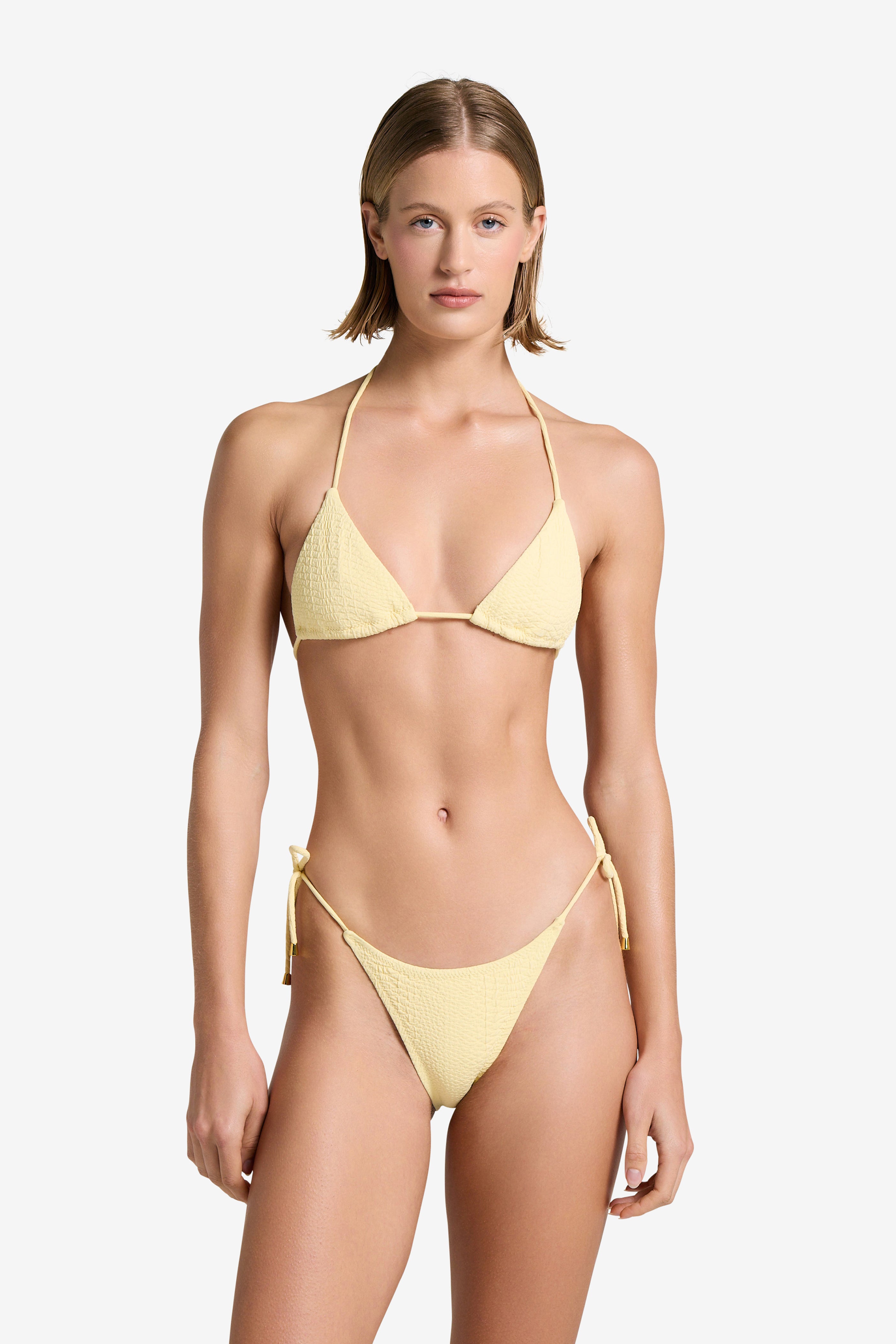 A person with short hair wears the Joni Top, a light yellow bikini from Gigi C Bikinis, featuring gold hardware. The textured fabric has string ties for an elegant touch, set against a plain white background that highlights the swimsuit.