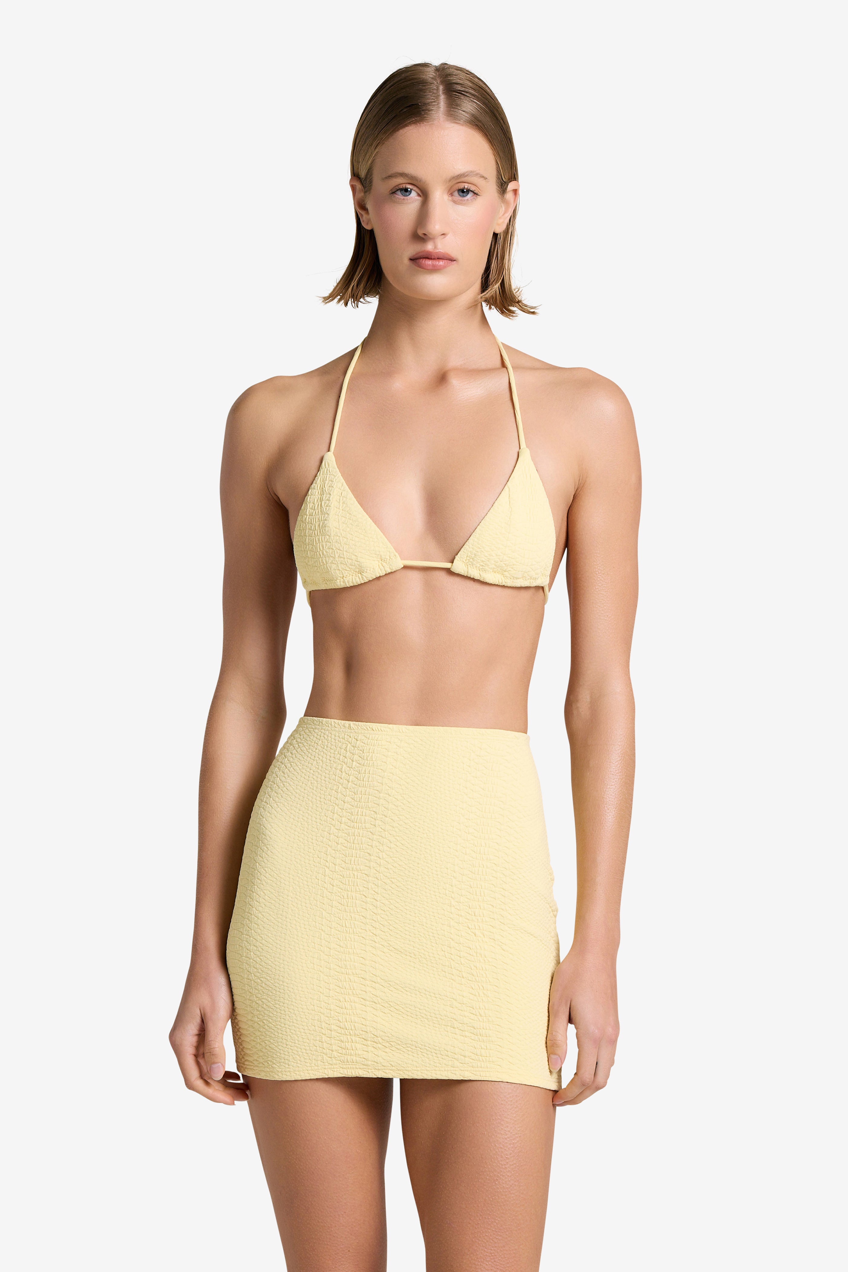 Against a plain white backdrop, the person wears a light yellow bikini-style top and Gigi C Bikinis Joelle Skirt in embossed fabric. The high-waisted skirt complements their short, straight hair, adding elegance to the look.