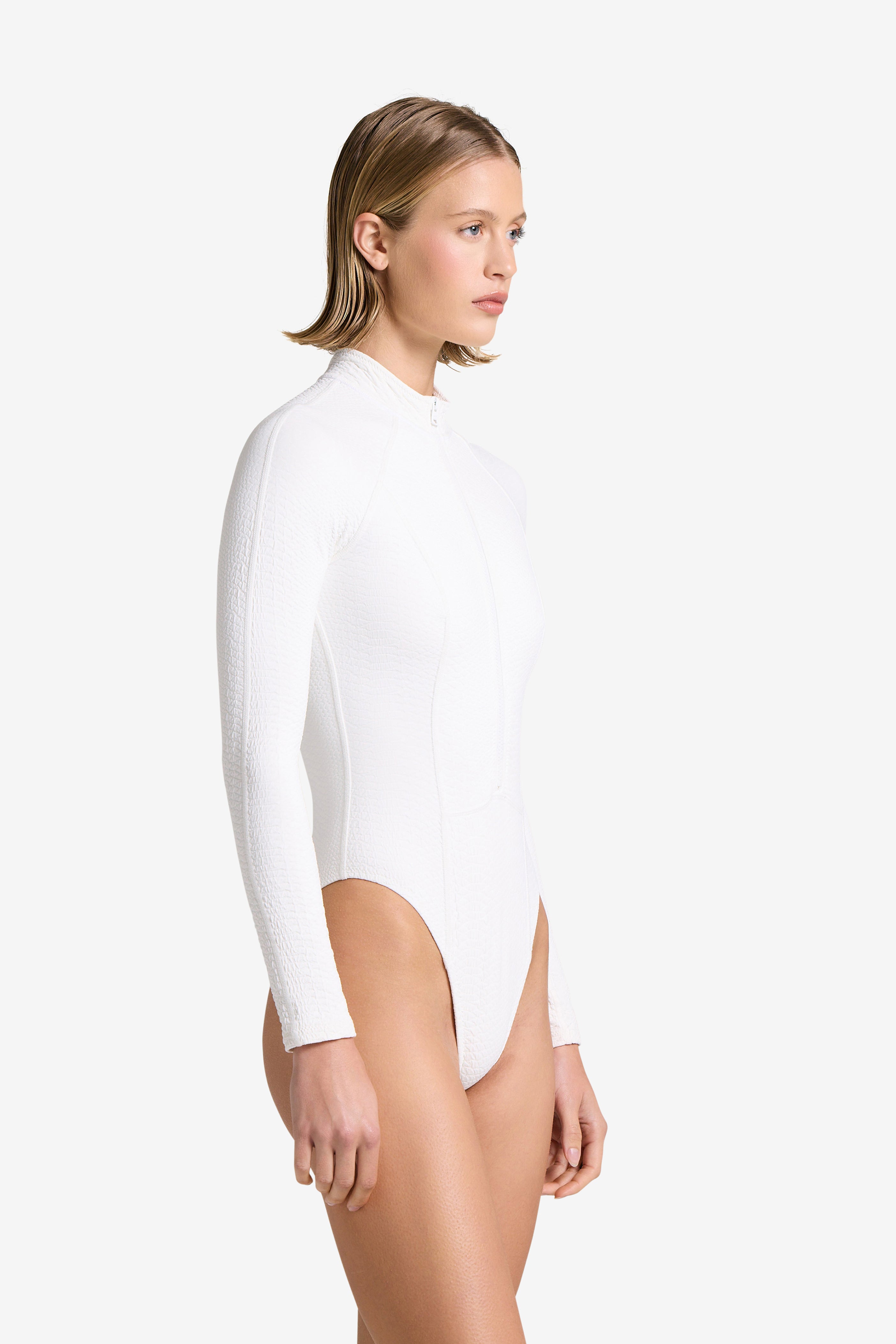 A person with short hair is modeling the Gigi C Bikinis Sophie Surfsuit in profile against a plain white background. This textured, luxe, faux snake fabric surfsuit features long sleeves, a white zip-up design, and a high-cut leg.