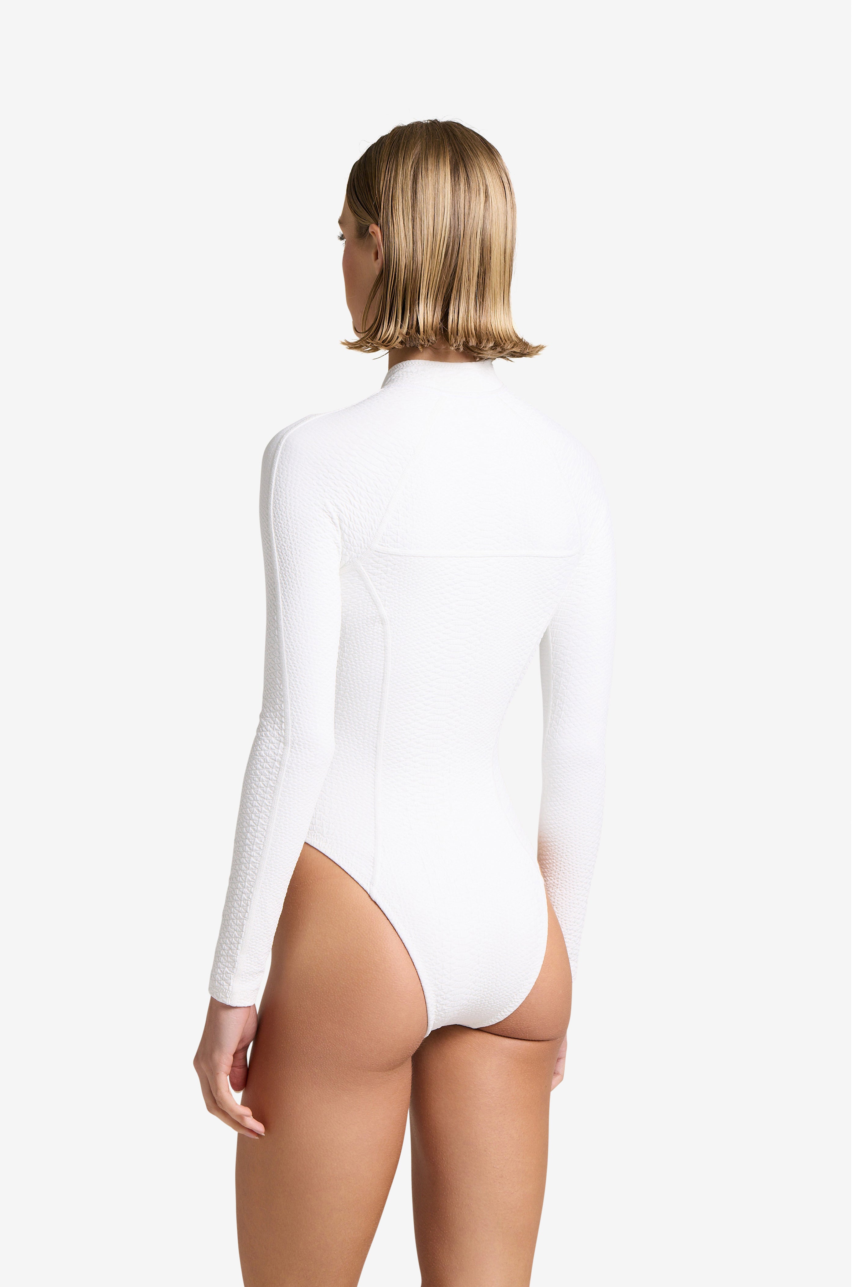 A person with short blonde hair poses against a plain white background in the Sophie Surfsuit by Gigi C Bikinis. The luxe surfsuit has a faux snake texture, featuring long sleeves and a high-cut leg, showcasing elegance and style from the back view.