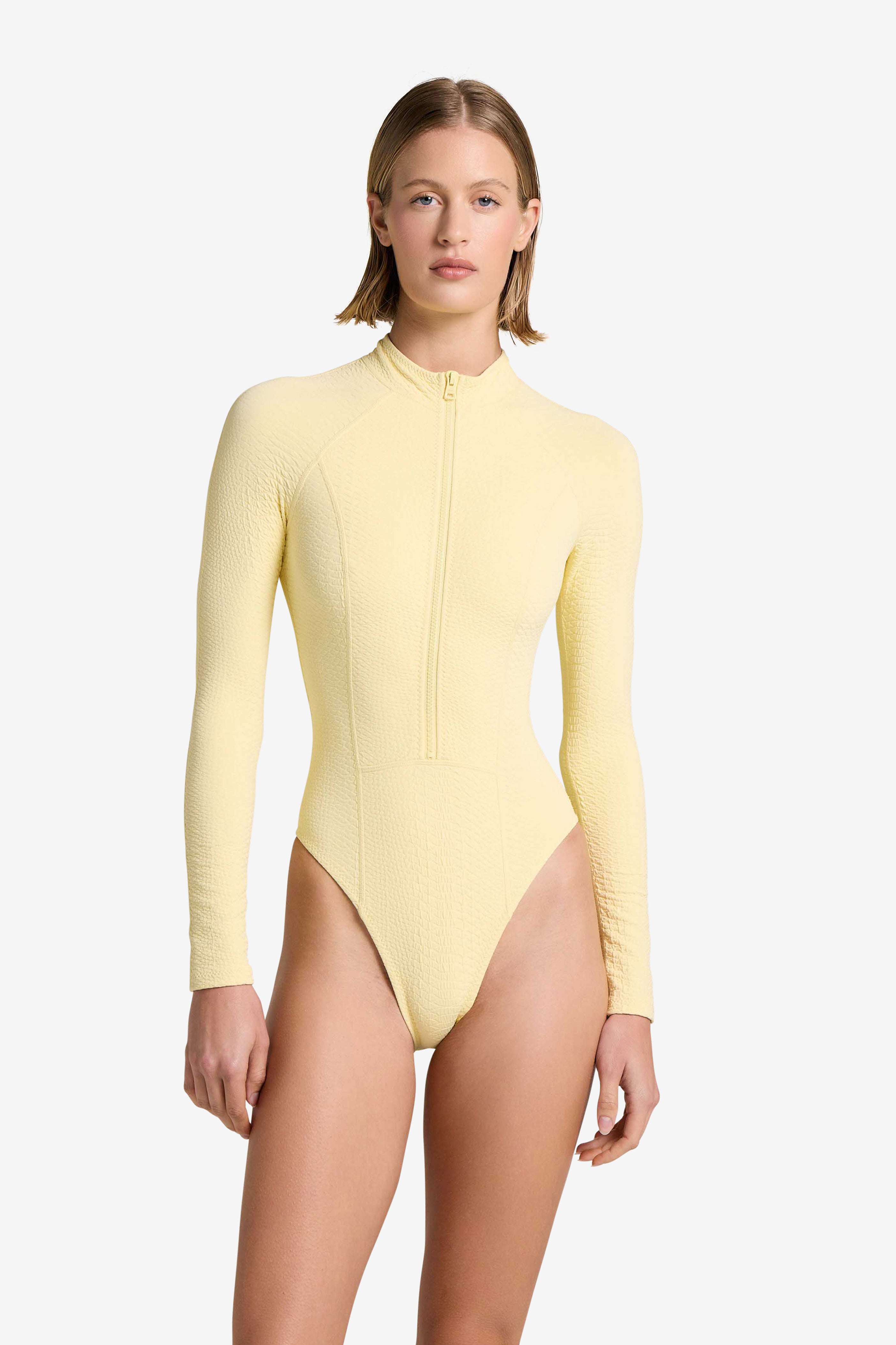 A person in a light yellow Sophie Surfsuit by Gigi C Bikinis, crafted from textured, snakeskin-embossed fabric with a front zipper, stands against a plain white background.