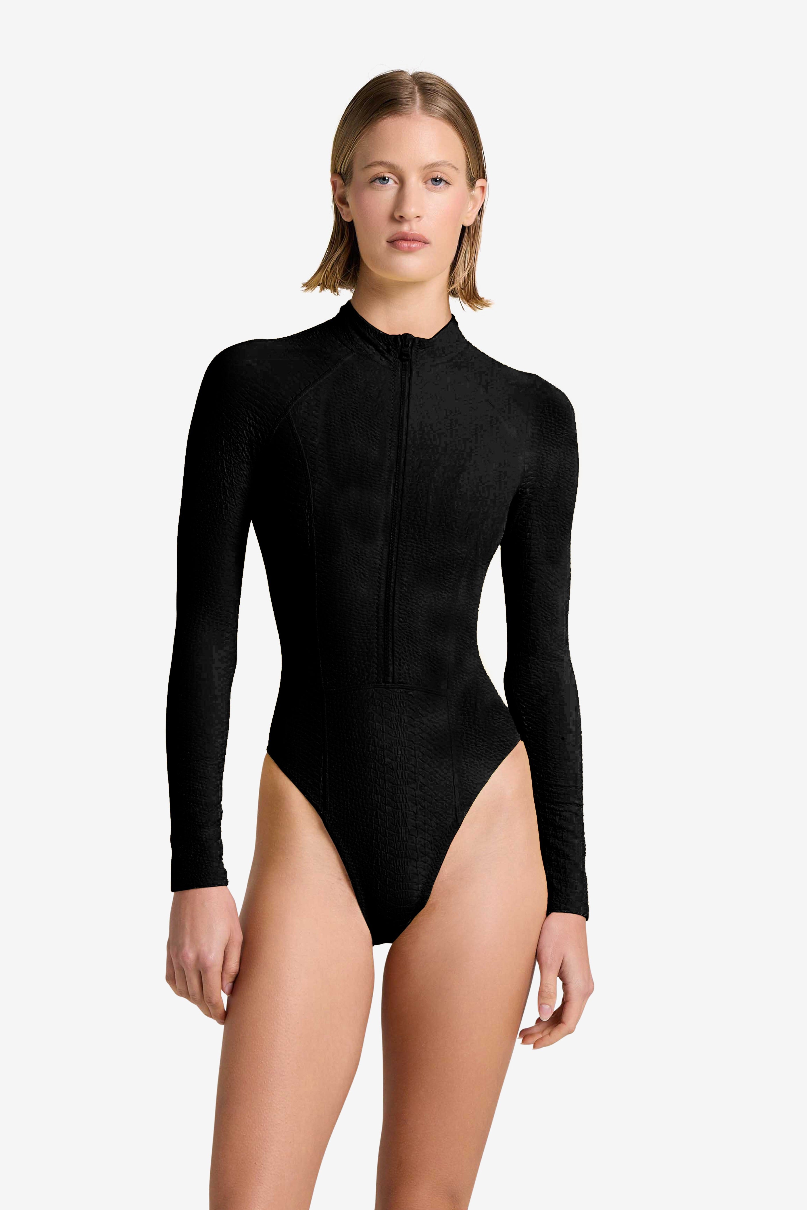 A person in a black, long-sleeve Sophie Surfsuit by Gigi C Bikinis stands against a white background. The surfsuit has subtle snakeskin embossed fabric and a front zipper. With short hair and a neutral expression, theyre ready for any water sport adventure.