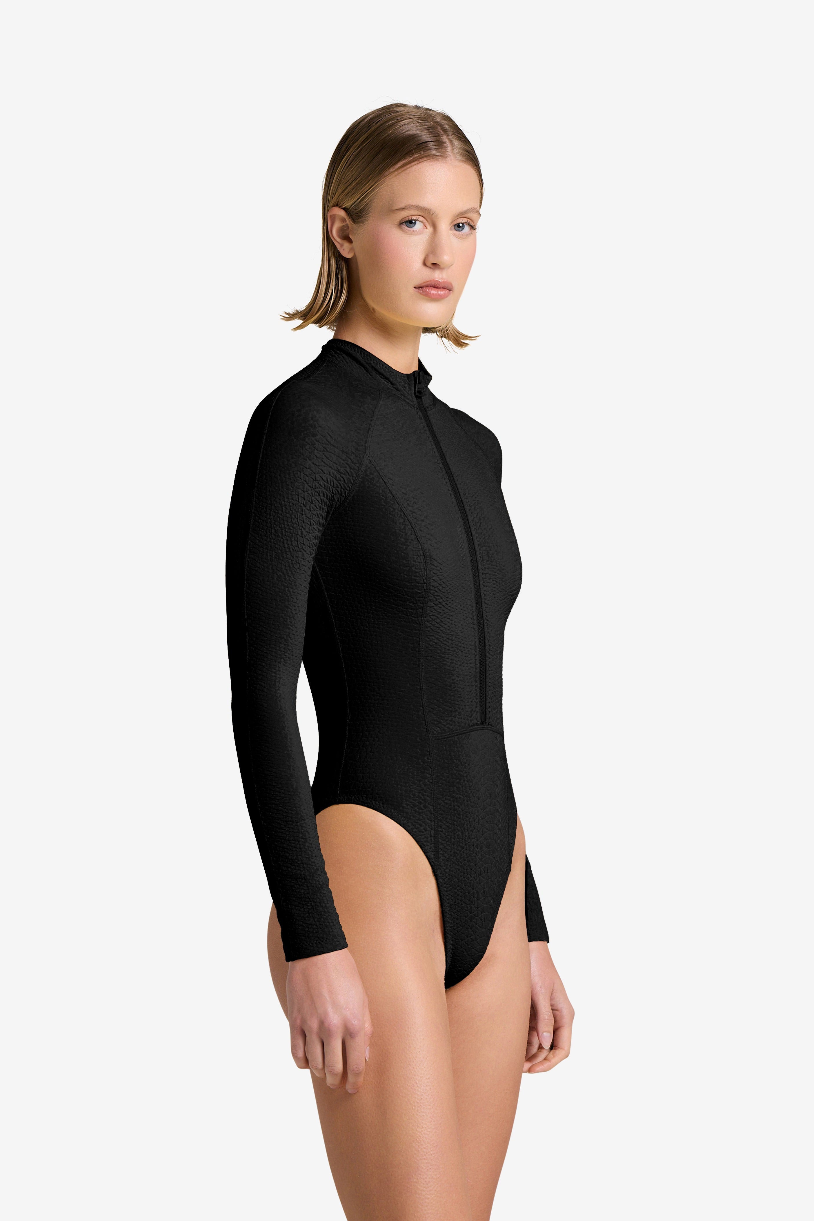 With short hair, they wear a long-sleeved, black bodysuit in snakeskin-embossed fabric. Standing against a white backdrop, theyre slightly turned and gazing at the camera, echoing the sleek design of Gigi C Bikinis Sophie Surfsuit.