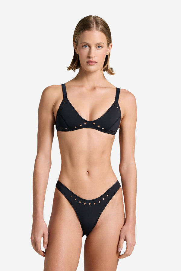 A person in a black Georgia Top bikini by Gigi C Bikinis, featuring laser cut detailing and small triangular cutouts, stands against a plain white background. They have short hair and a neutral expression.