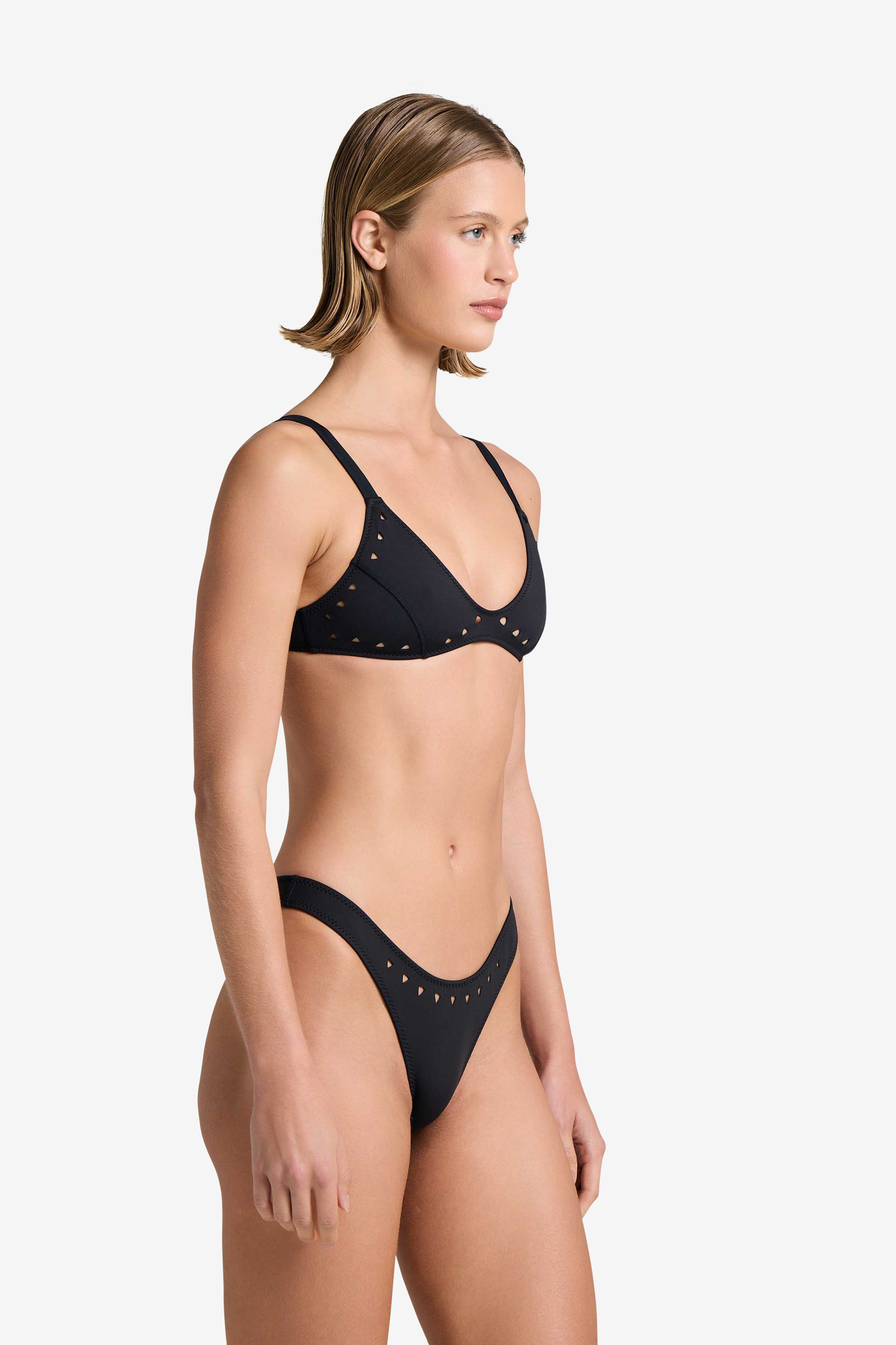 Someone with short hair wears the Georgia Top by Gigi C Bikinis, which features black laser cut detailing, as they stand in profile against a plain white background.