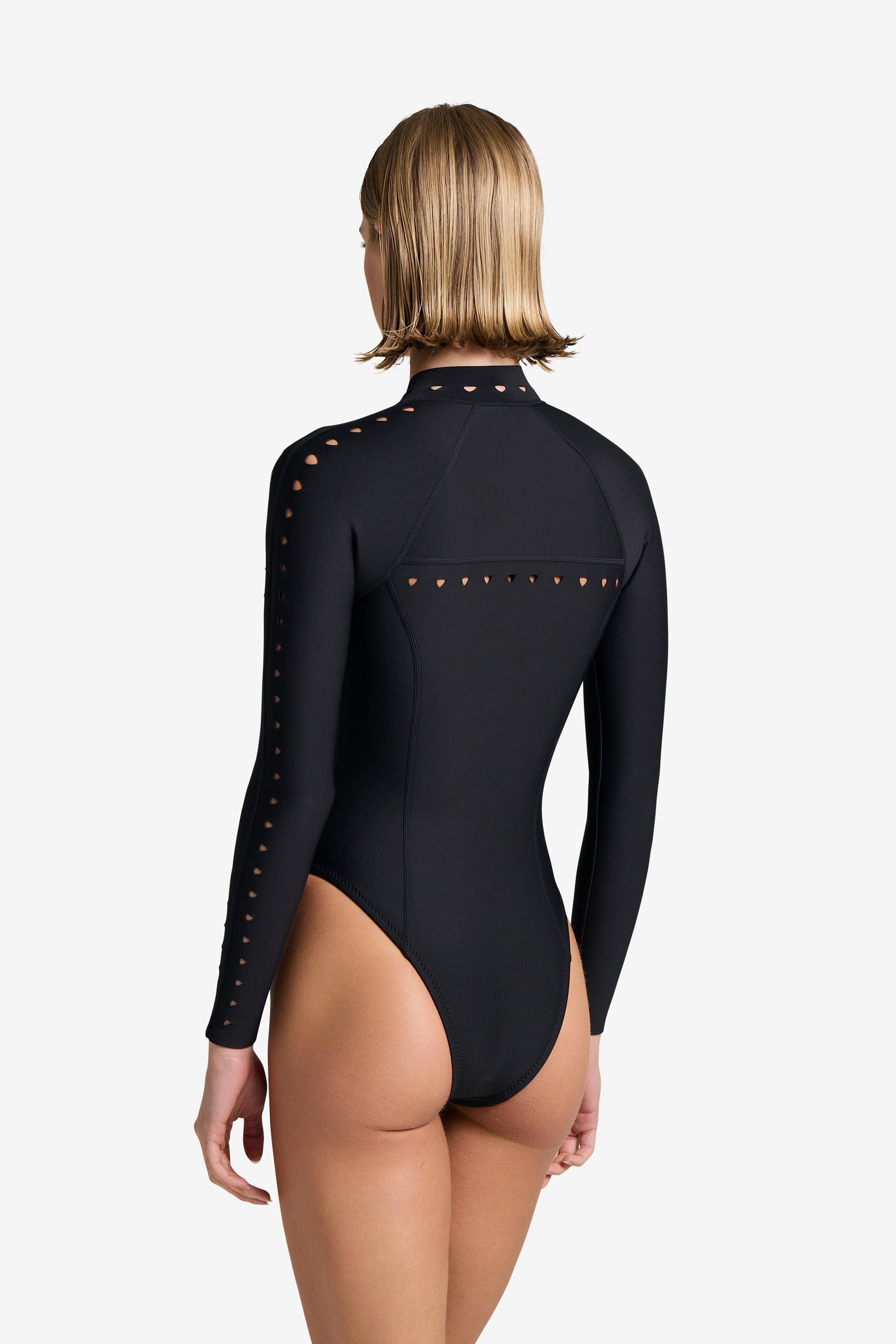 A person with short blonde hair is wearing the Riley Surfsuit by Gigi C Bikinis. This black, long-sleeve, high-neck swimsuit is made from scuba fabric and features laser cutouts on the sleeves and back as they pose with their back to a plain white camera background.