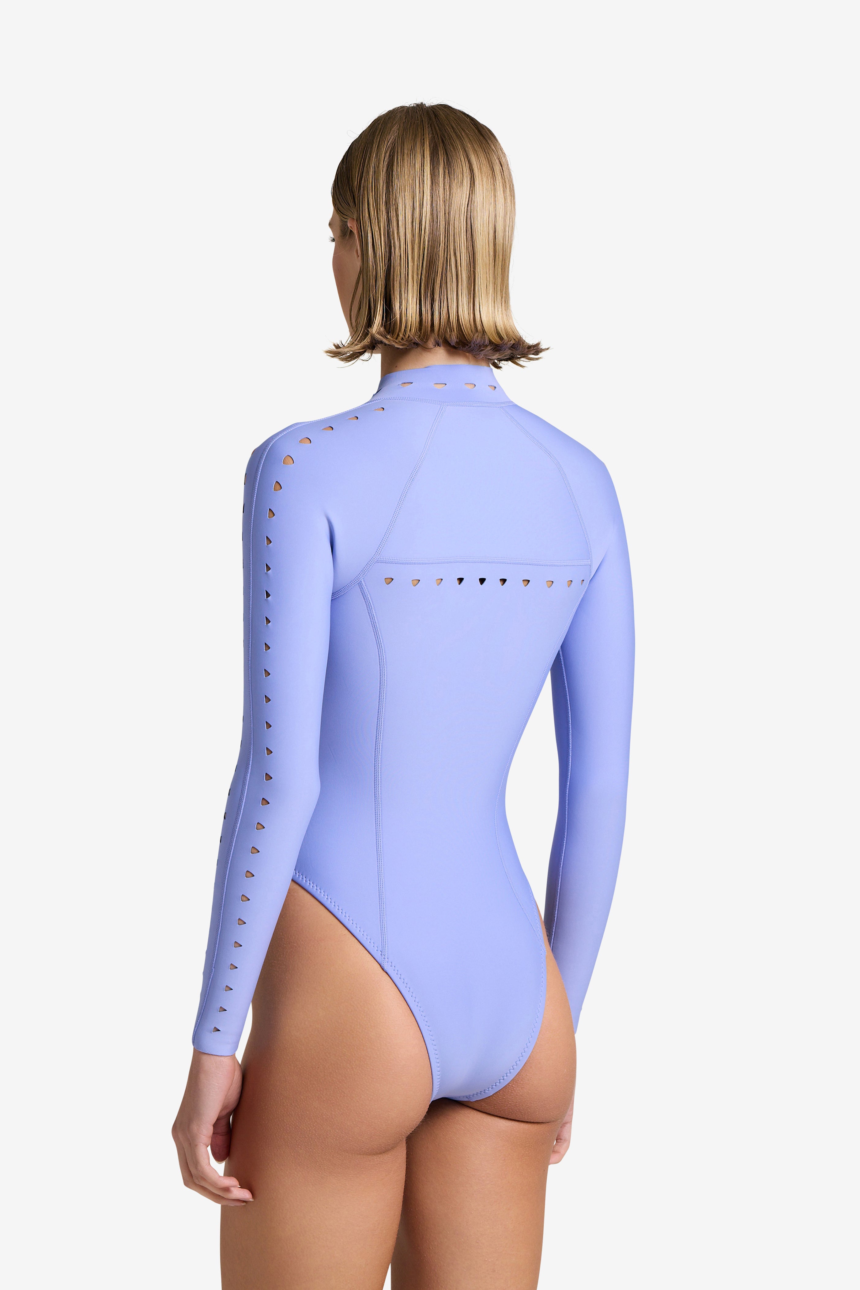 From behind, a person with short blonde hair wears the Riley Surfsuit by Gigi C Bikinis—a long-sleeve, light purple scuba fabric swimsuit featuring a front zip closure and triangle laser cutouts on the sleeves and upper back, all set against a plain white backdrop.