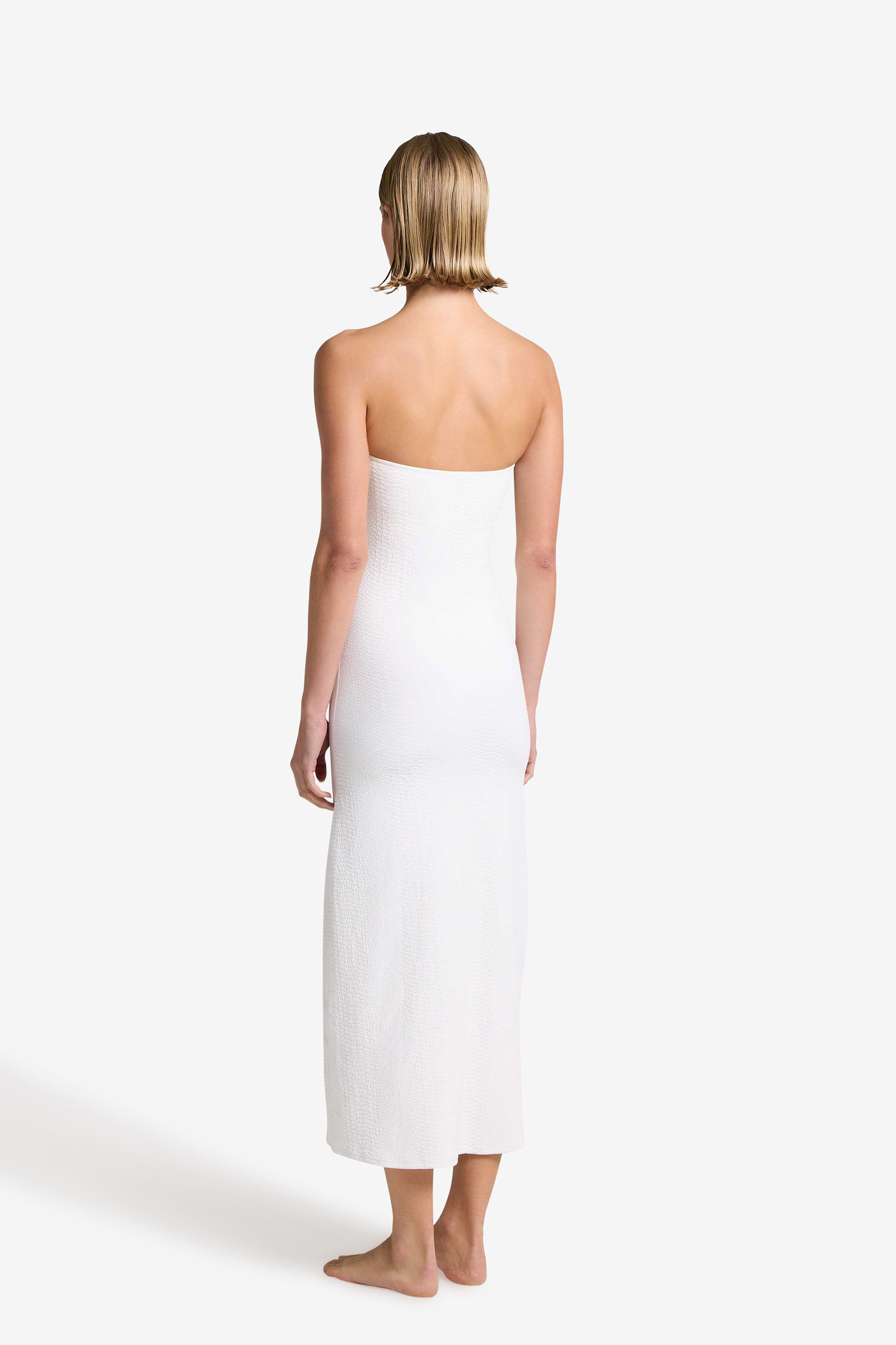 A person with short hair, facing away, wears the Maya Dress by Gigi C Bikinis. The elegant strapless ivory design, crafted from textured fabric, reaches their ankles as they stand barefoot against a plain white background.