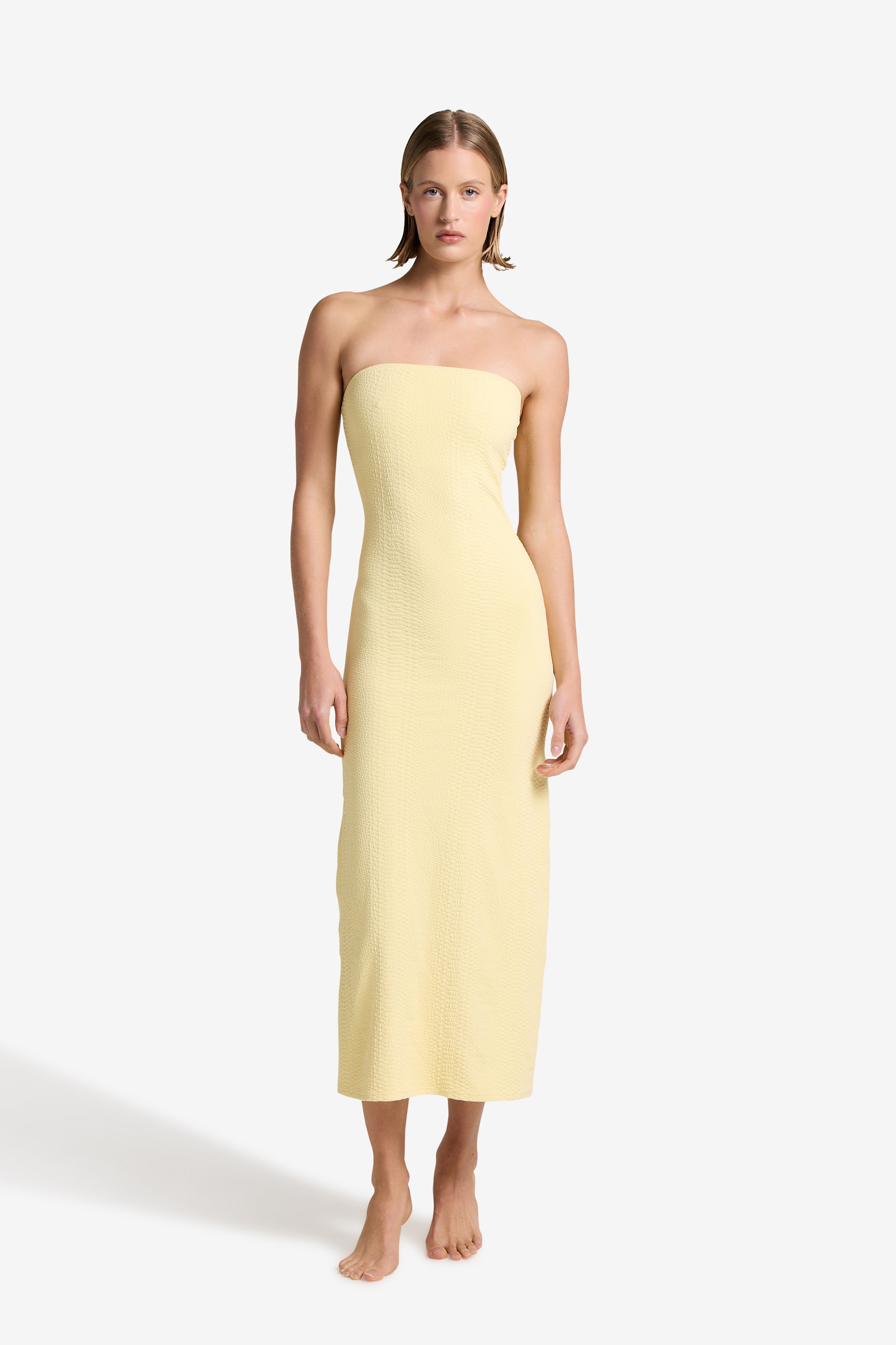 Barefoot against a plain backdrop, a person wears Gigi C Bikinis elegant Maya Dress in light yellow. The strapless, ankle-length design complements their relaxed posture as short, straight hair frames their serene demeanor, with arms at their sides.