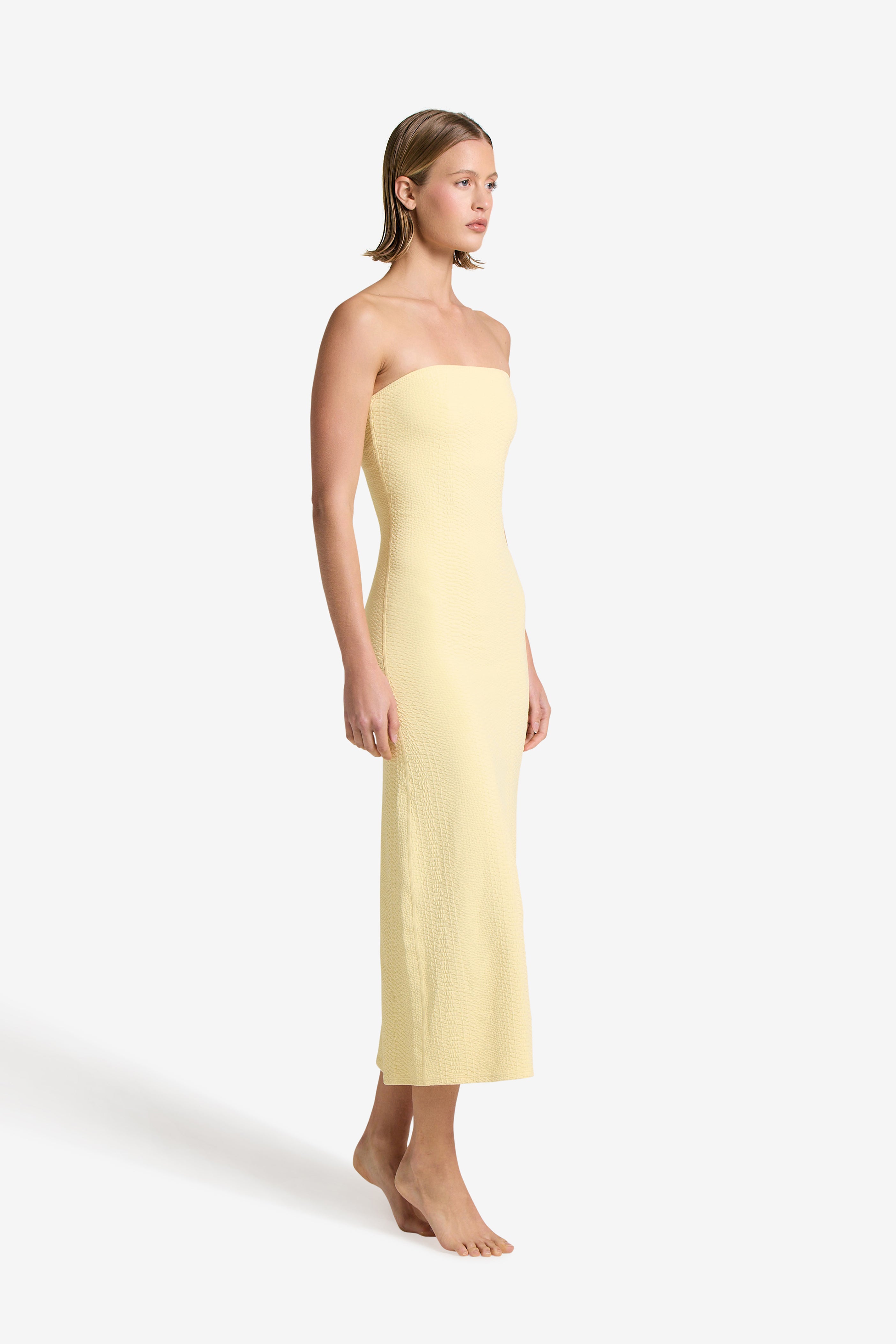 A woman stands barefoot against a light background, wearing a yellow ankle-length Maya Dress by Gigi C Bikinis. Facing slightly left with relaxed arms, her short, neatly styled hair enhances an elegant look.
