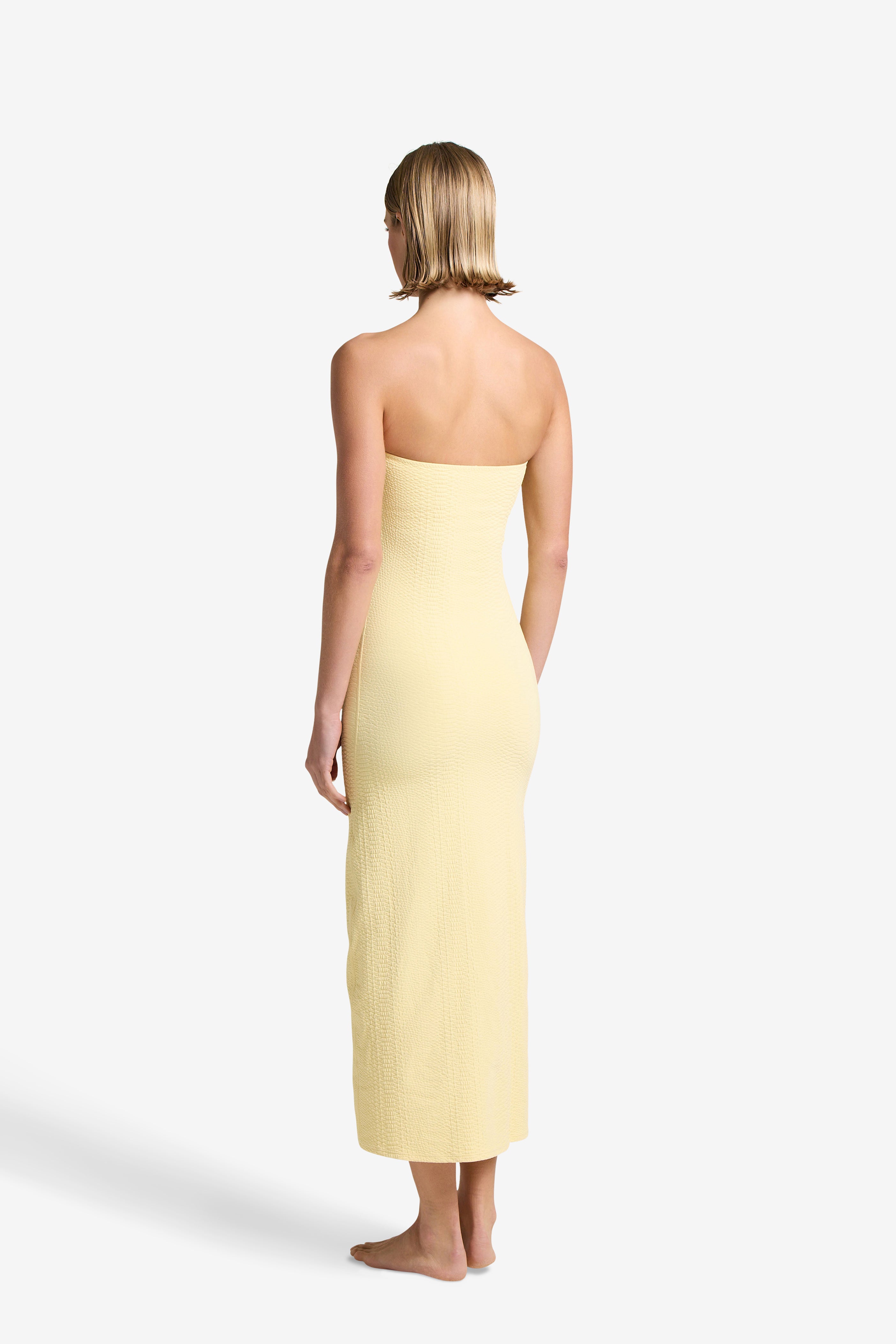 A person with short hair, facing away, wears the Maya Dress by Gigi C Bikinis. The elegant strapless yellow design, crafted from textured fabric, reaches their ankles as they stand barefoot against a plain white background.