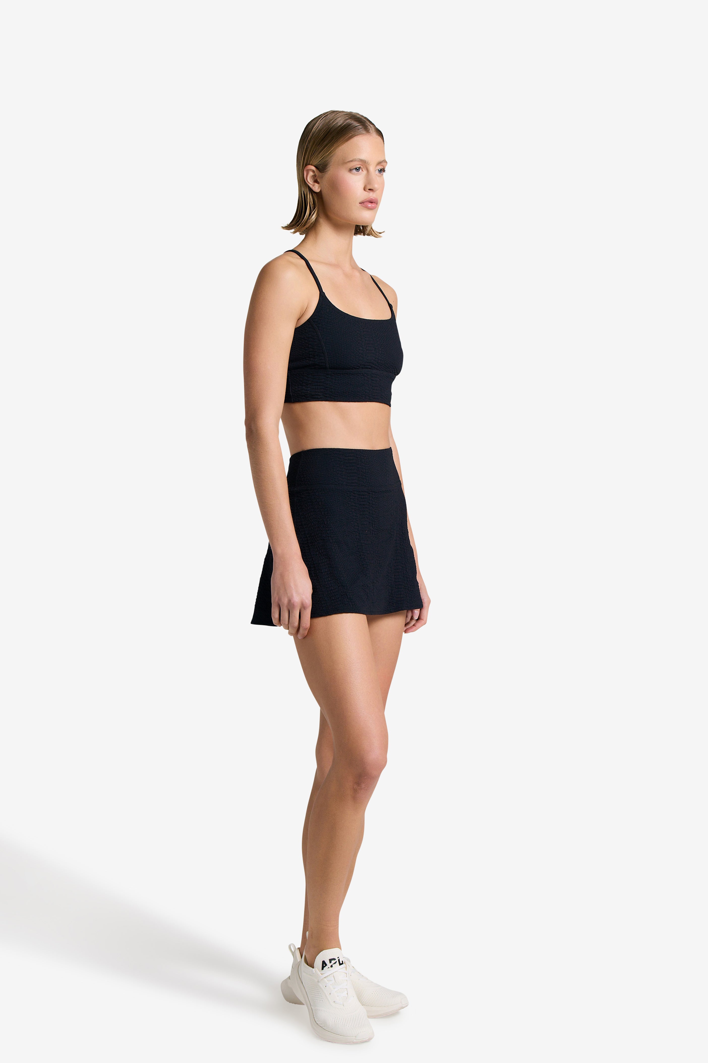 A person stands against a white background wearing the Gigi C Sport Heidi Skort in black bi-stretch compression waffle jacquard fabric, paired with a matching sports bra and white sneakers. They face right with a neutral expression and arms relaxed by their sides.