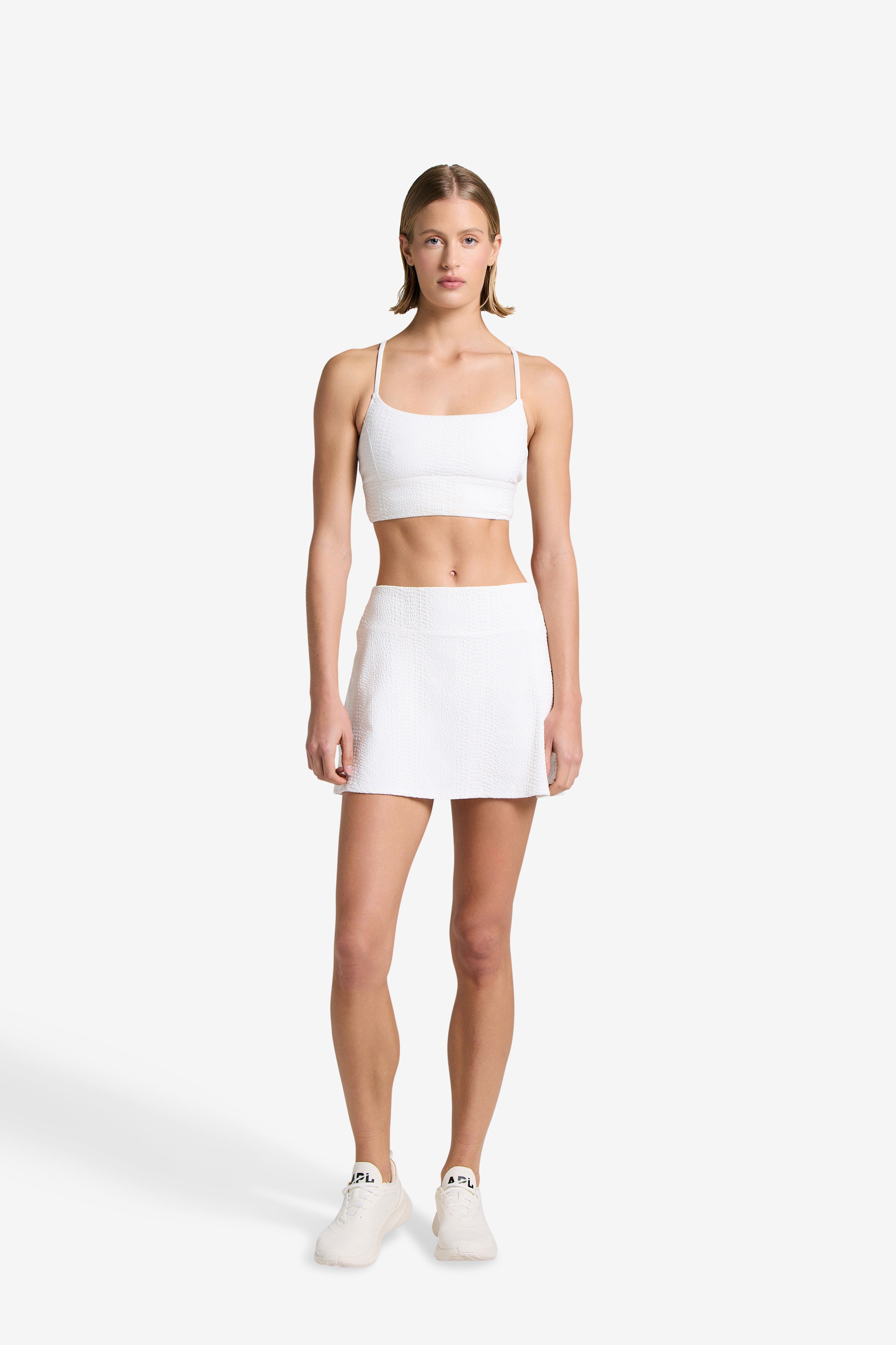 A woman in a white athletic outfit poses against a plain background, highlighting the Finle Bra from Gigi C Sport. She pairs it with a skort, wearing sneakers and featuring middle-parted hair and a neutral expression, with subtle snakeskin-embossed fabric accentuating elegance.