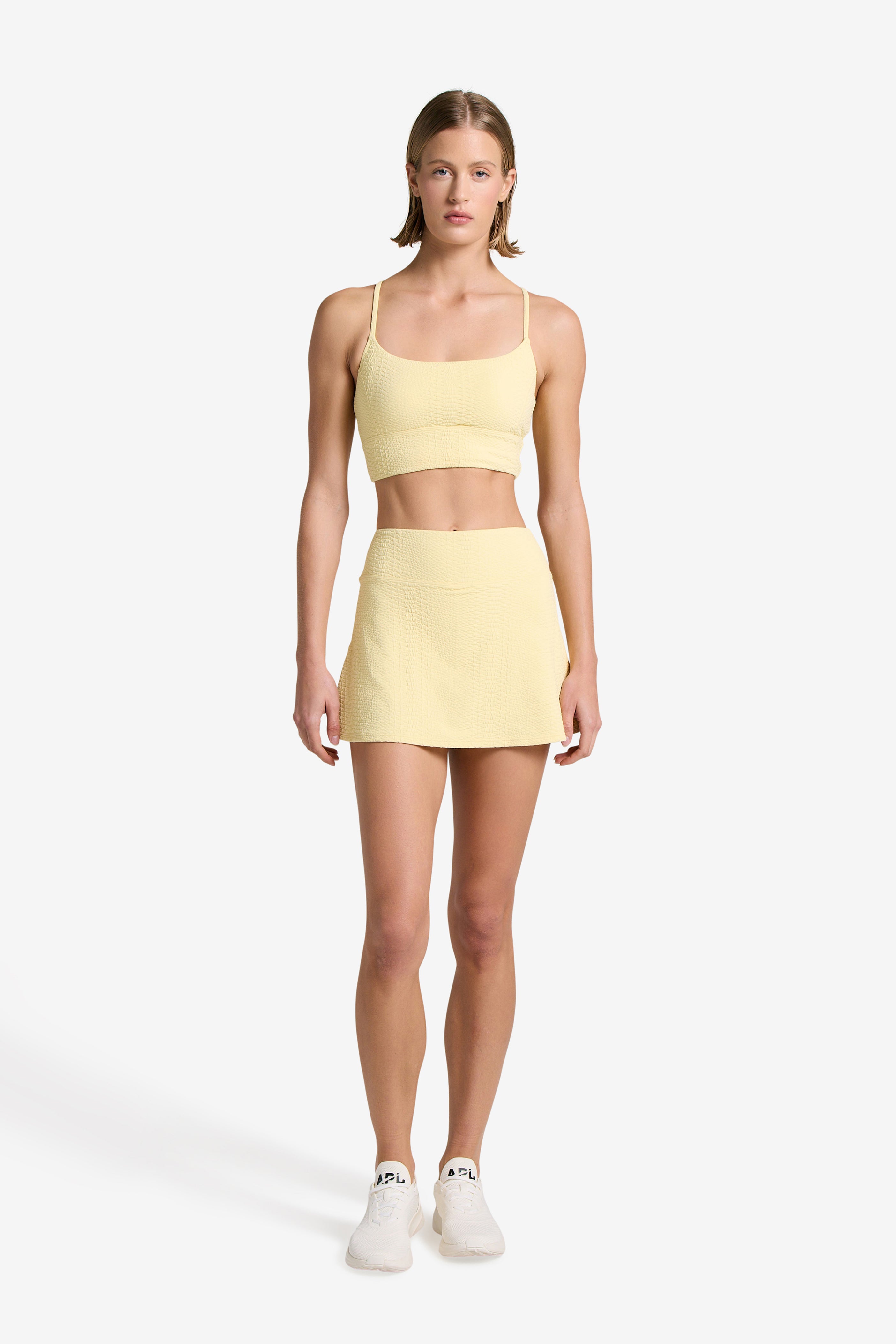 A person stands against a plain white background, wearing a yellow Finle Bra and matching Heidi Skort by Gigi C Sport in snakeskin-embossed fabric. They have short hair, wear white sneakers, and keep their hands relaxed by their sides.