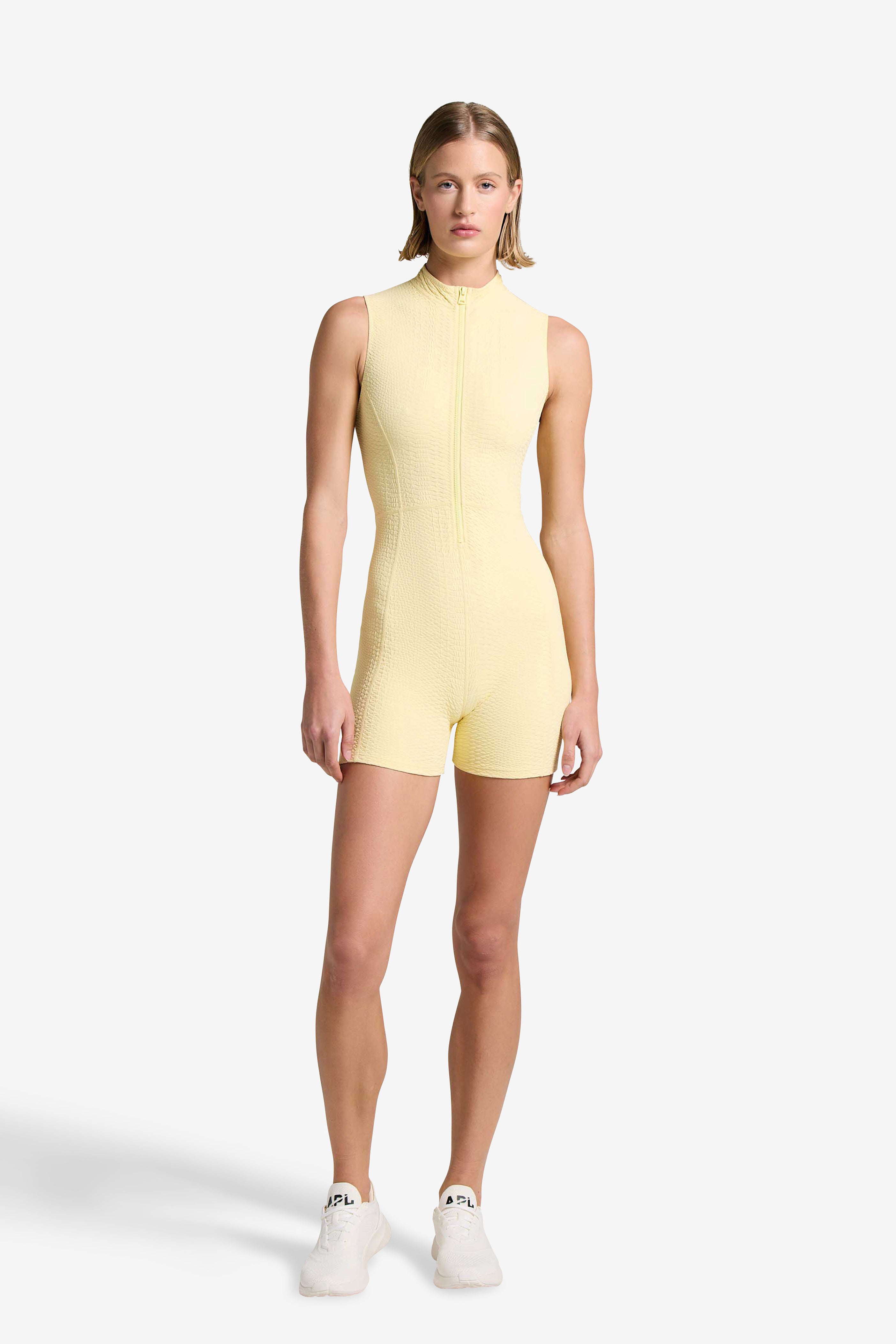 A person stands against a plain white background, wearing a sleeveless yellow Gigi C Sport Keelan Romper and white sneakers. With short hair and a neutral expression, they face the camera to highlight the rompers customizable coverage.