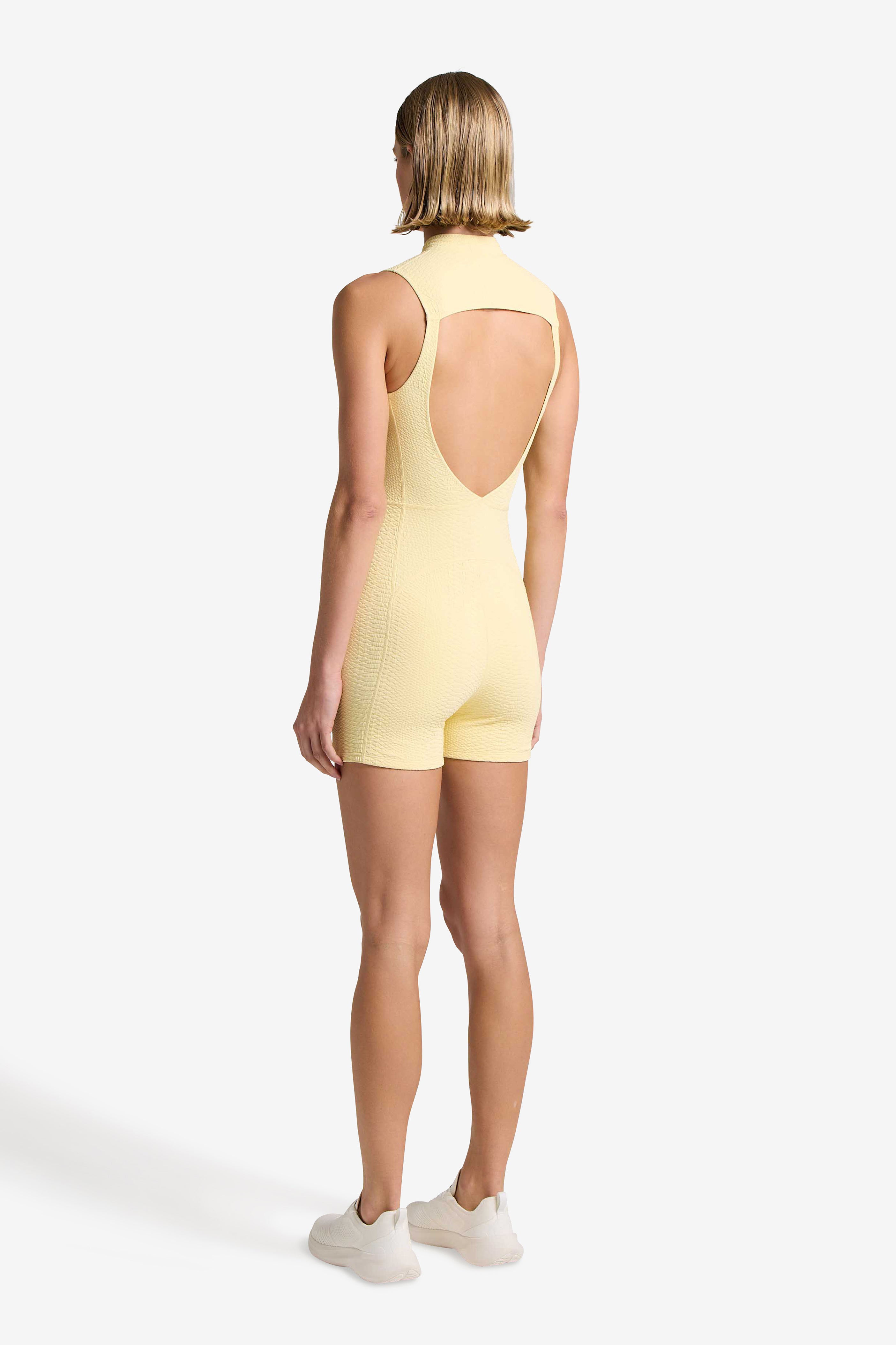 A person faces away from the camera, highlighting the sleeveless, open-back yellow Keelan Romper by Gigi C Sport with white sneakers. The plain backdrop enhances the rompers sleek design and striking silhouette.