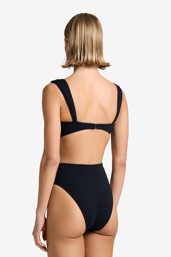 A person with short blonde hair is shown from behind, wearing the Dawson Top by Gigi C Bikinis. The textured black bikini is made from snakeskin-embossed fabric, featuring wide straps connecting at the back and high-waisted bottoms against a plain, light-colored background.