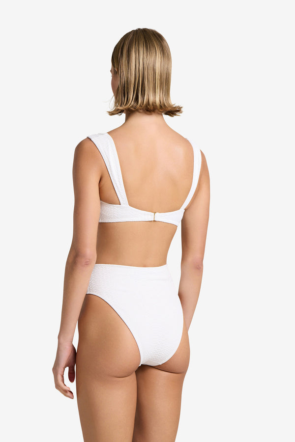 A woman with short blonde hair models the Melinda Bottom, a textured white two-piece swimsuit by Gigi C Bikinis in snakeskin-embossed fabric. The top has wide straps and a back clasp. She stands against a plain white background, highlighting her chic swimwear.