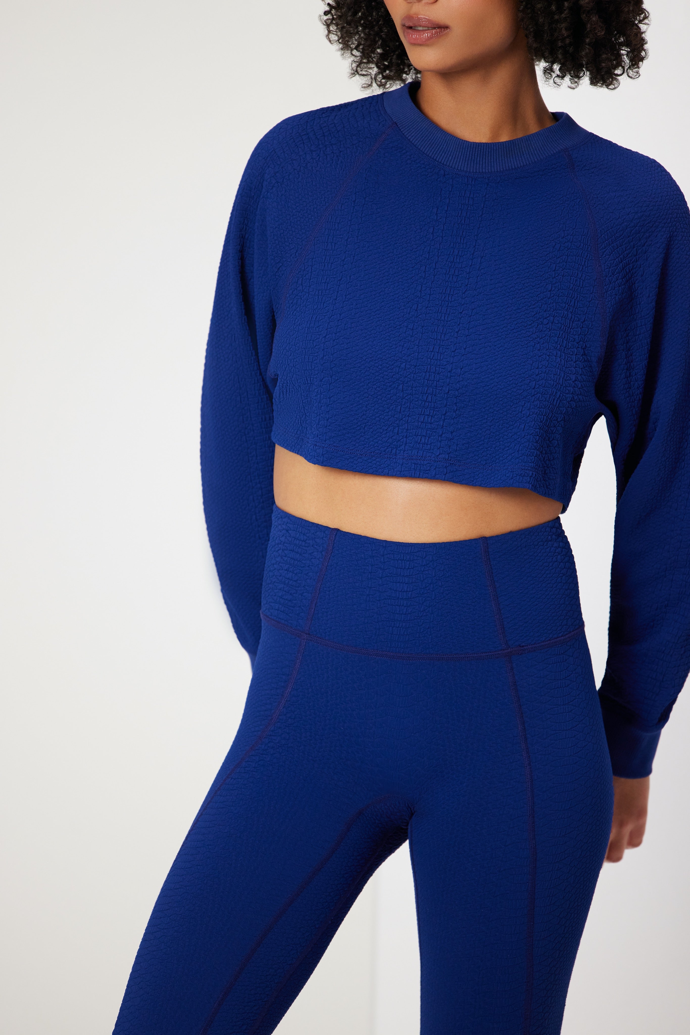 Wearing a cobalt blue Isabella Pullover from Gigi C Sport, a textured crop top with matching high-waisted leggings, enhances the seamless design against the plain white backdrop.