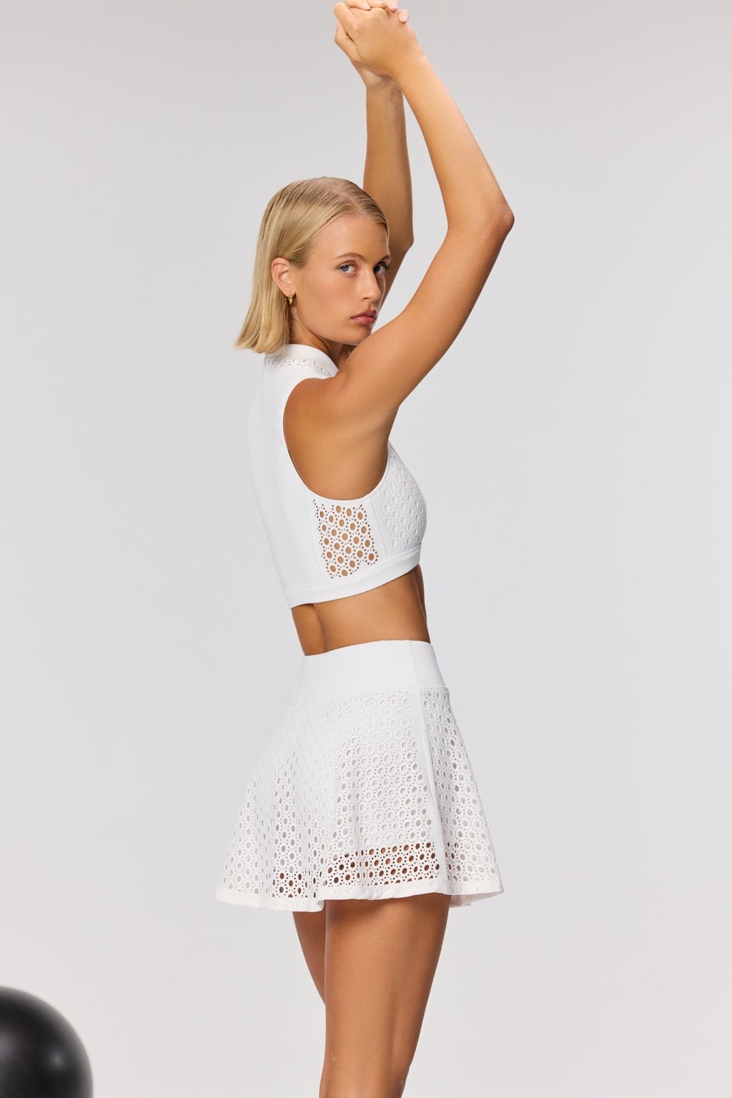 A person poses with raised arms, gazing sideways while wearing a white sleeveless Briar Bra Top from Gigi C Sport and a skirt. The outfit, made of sculpting sport fabric with intricate cutouts, stands out against the plain neutral background, highlighting its athleisure elegance.