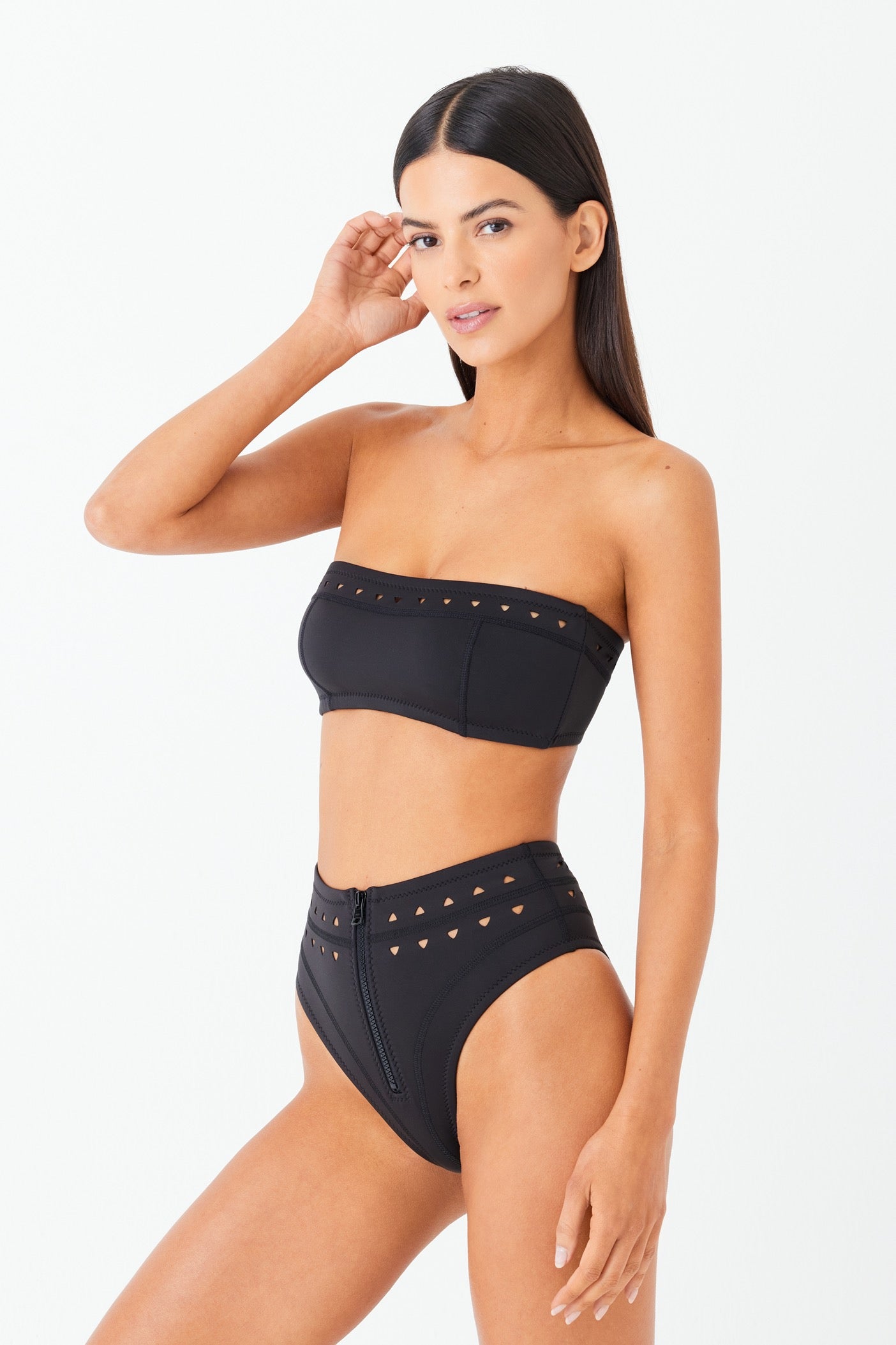 A woman models Gigi C Bikinis McKenzie Bottom, a black strapless bikini with laser triangle cutouts. She stands against a plain white background, looking towards the camera with one hand touching her head.