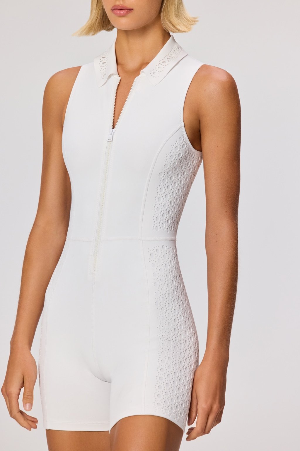 A woman stands confidently in a sleeveless white Mina Romper from Gigi C Sport, featuring eyelet side details. Her blonde bob adds style to the versatile activewear, enhancing its fashion and function against a plain background.