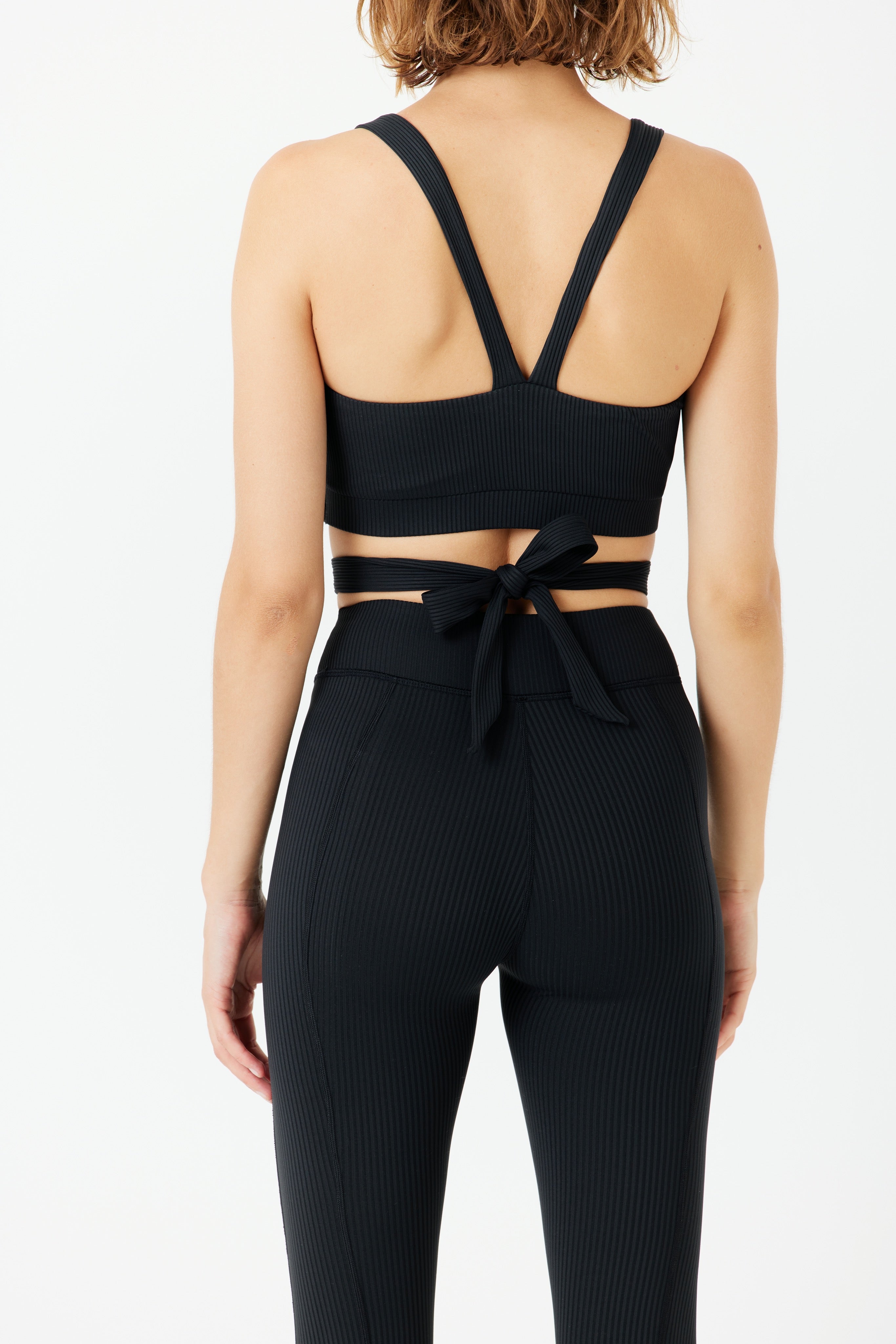 A person models the Phoebe high-waisted leggings by Gigi C Sport in a black textured athletic set, showcasing buttery soft power rib fabric. The ensemble includes a crisscross sports bra with a ribbon-tied waist, all against a plain white backdrop.