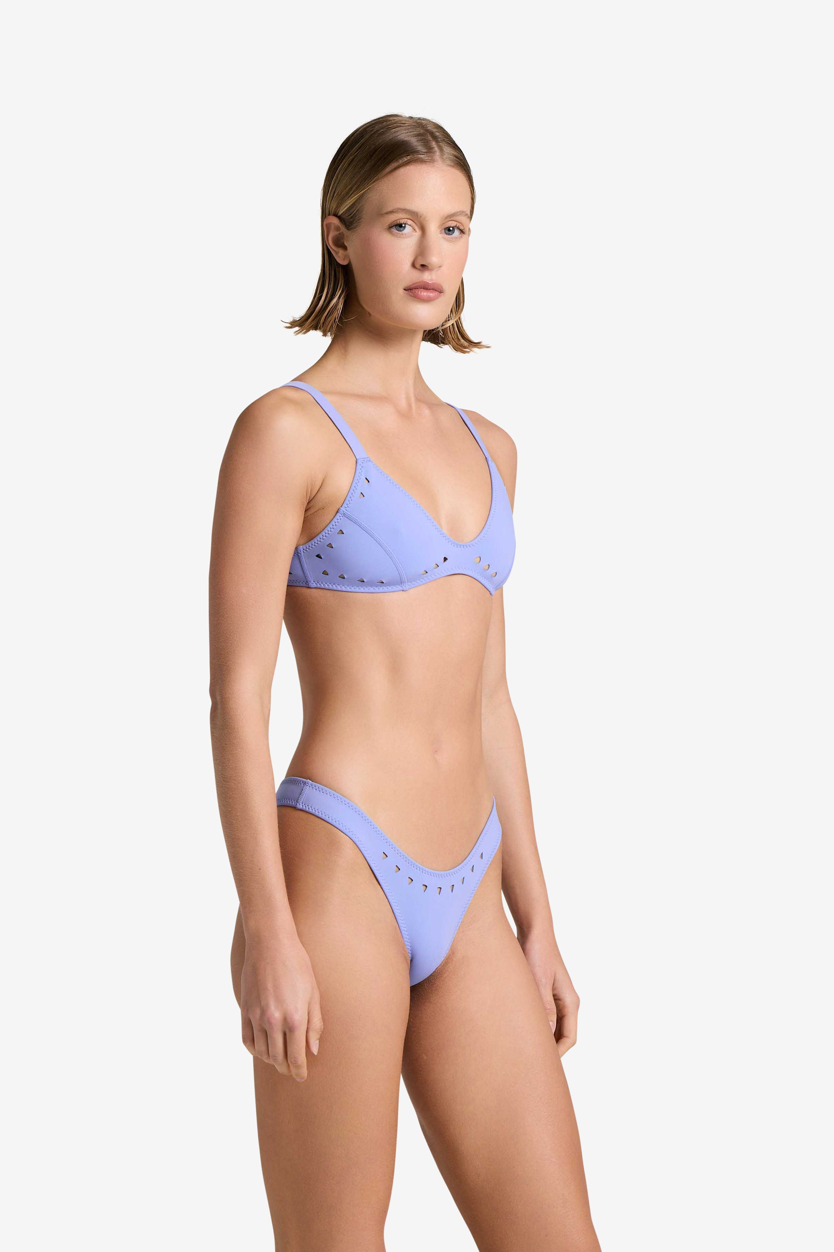 A woman with short hair wears the Georgia Top bikini by Gigi C Bikinis, featuring a light purple shade with laser cut detailing. She stands against a white background, looking directly at the camera. The bikini offers adjustable straps and high-cut bottoms for a modern twist on a classic style.