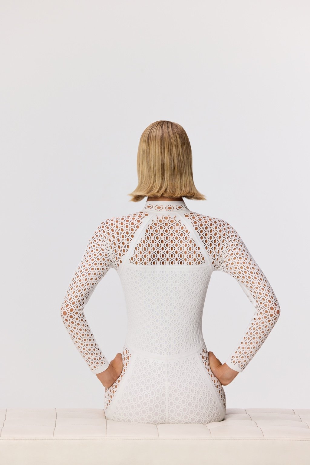 A blonde sits on a white surface, facing away, in a fitted Rowen Surfsuit by Gigi C Bikinis. The wax-coated scuba fabric and high neckline add elegance, while their sleek bob enhances the chic style against the plain backdrop.
