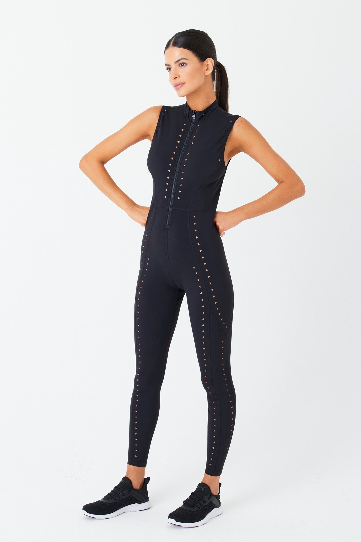 Model poses confidently against a white backdrop, flaunting the Gigi C Sport Sarah Jumpsuit in black LYCRA with a zipper front, intricate laser cutout designs, and dotted patterns. They have long hair tied back and wear black athletic shoes.