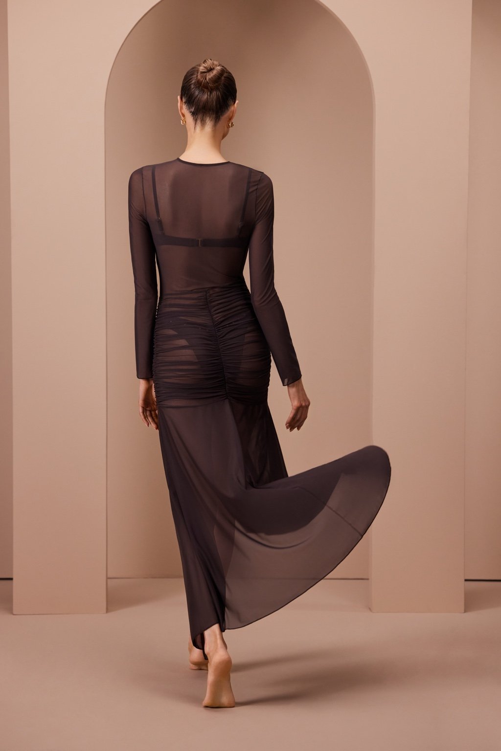 A person with a hair bun wears the Gisele Maxi by Gigi C Bikinis, made from silky mesh fabric, standing in an arch-filled minimalist setting. They look right with one foot slightly raised, creating an elegant sense of motion in the scene.