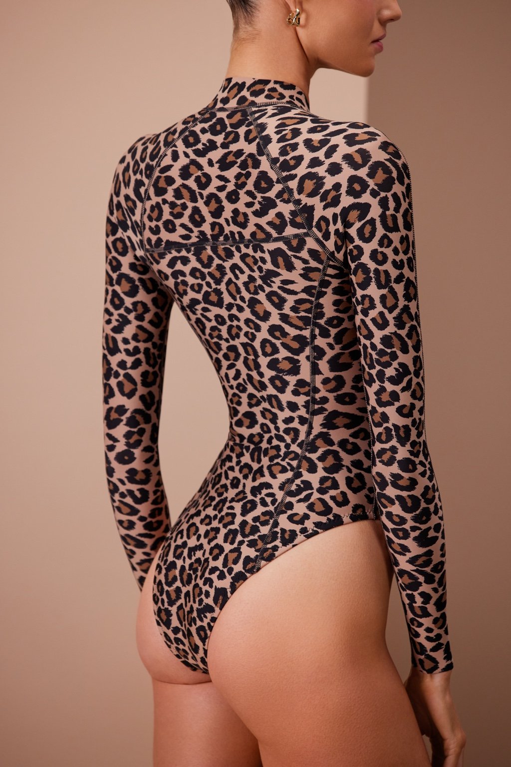 A person poses with their back to the camera in the Kendall Surfsuit by Gigi C Bikinis, a long-sleeve leopard print bodysuit made from signature scuba fabric, set against a neutral background.