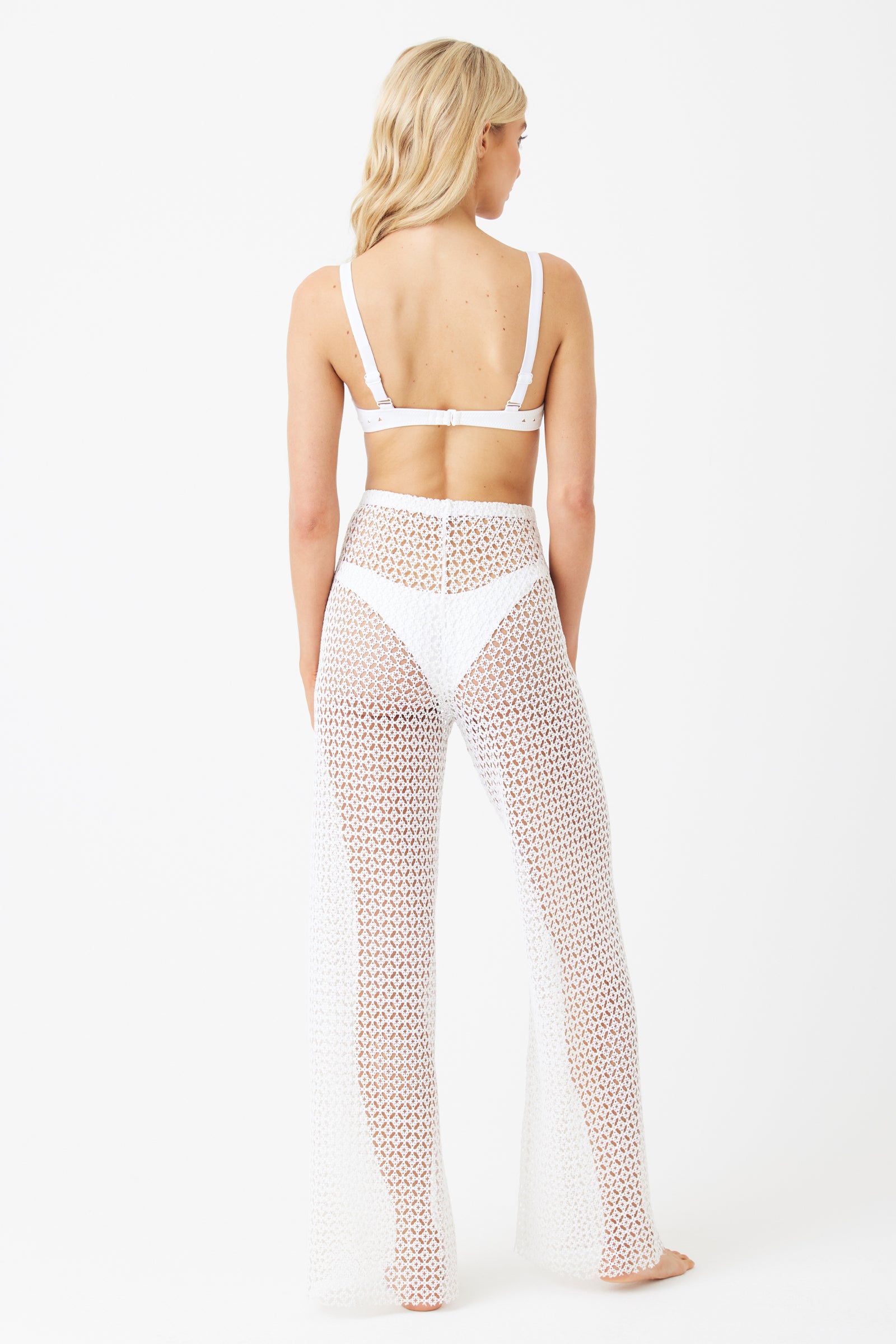 A person with long blonde hair is facing away, wearing a white bra top and sheer Andy Pant by Gigi C Bikinis, crafted from diamond lace fabric in a circular pattern. The background is plain white.