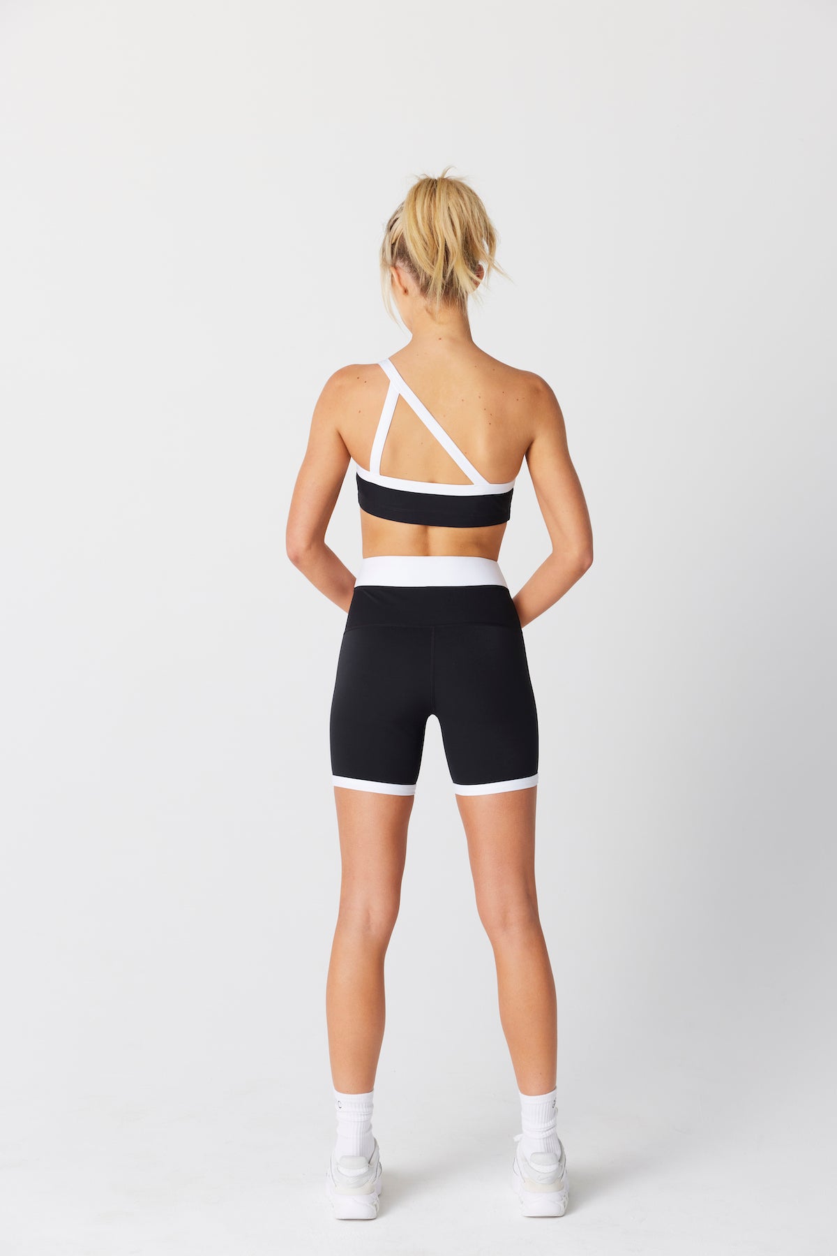 A woman stands with her back turned, wearing Gigi C Sports black and white Addie Asymmetric Bra with matching shorts. Her hair is styled in a ponytail, complemented by sleek white sneakers. The breathable knit fabric offers quick-drying comfort against the plain white background.