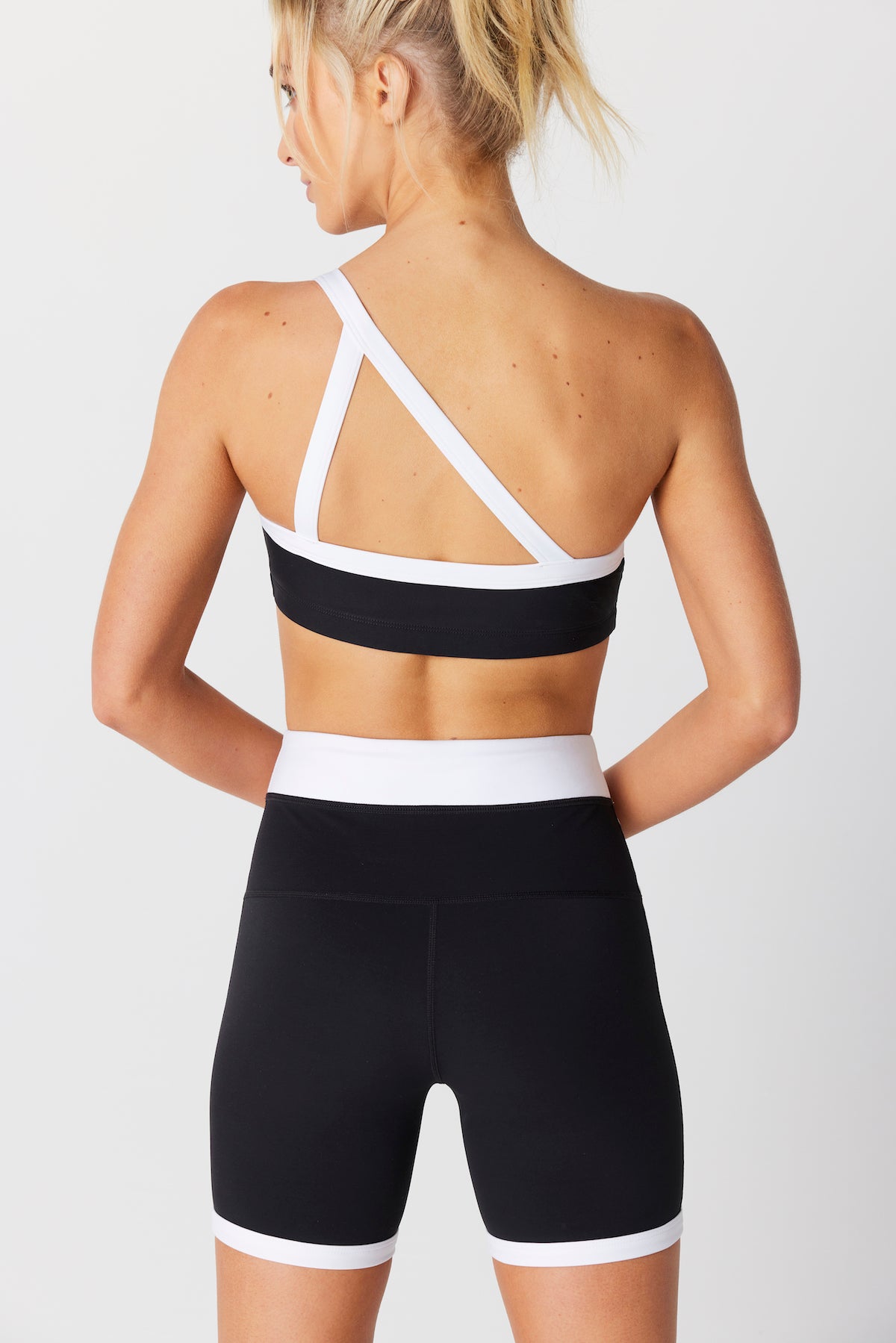 A blonde-haired person in a ponytail wears the Gigi C Sport Addie Asymmetric Bra and matching black shorts with white trim. The bra features cross-back straps, the shorts have a wide waistband, and both are perfect for active days. They pose against a white background with their back to the camera.
