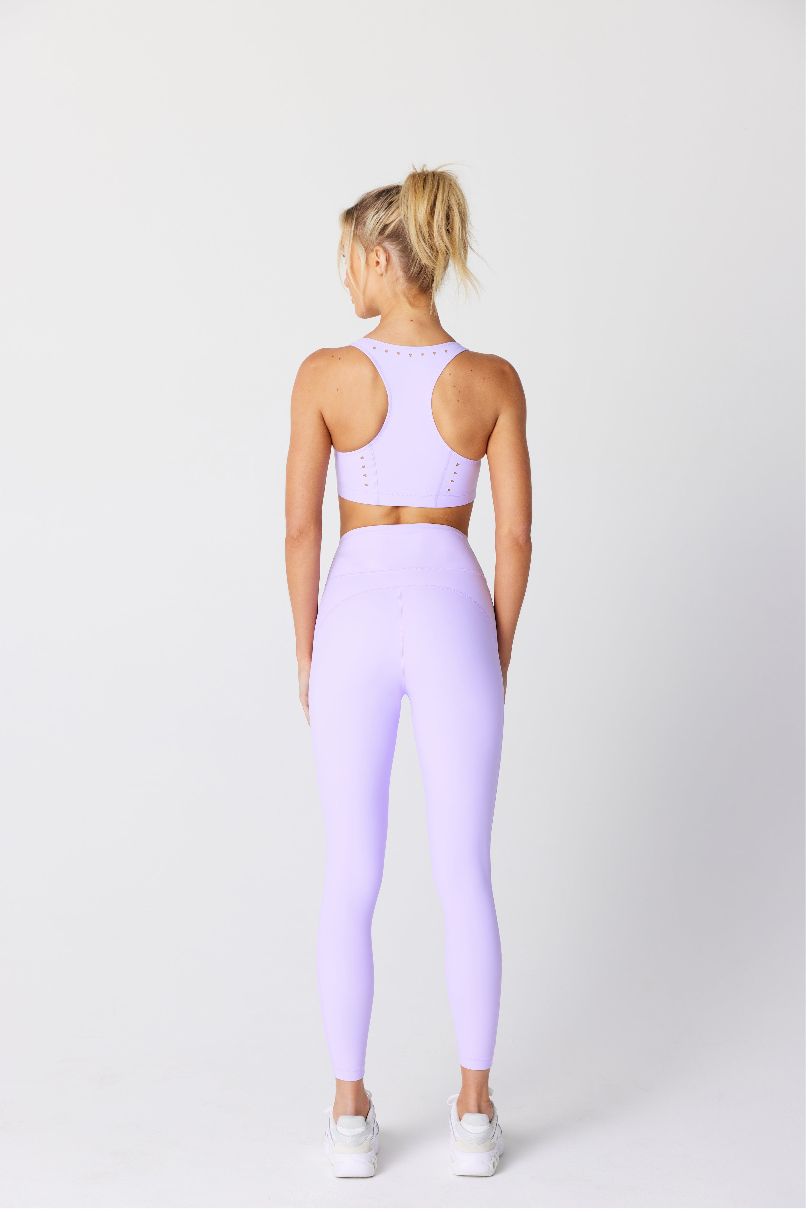 A woman models the Gigi C Sport Annabelle Bra Top in lavender, made from recycled bonded jersey. It features removable pads and is paired with leggings. Her high ponytail and white sneakers complete the look against a white background.