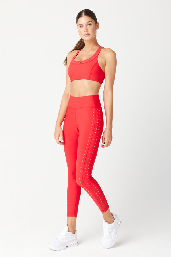 A woman poses against a plain background in a matching high-waisted red sports bra and Gigi C Sports Katrina Legging set with white dot accents. Her long hair is tied back, and shes wearing white sneakers, projecting sleek style and comfort.