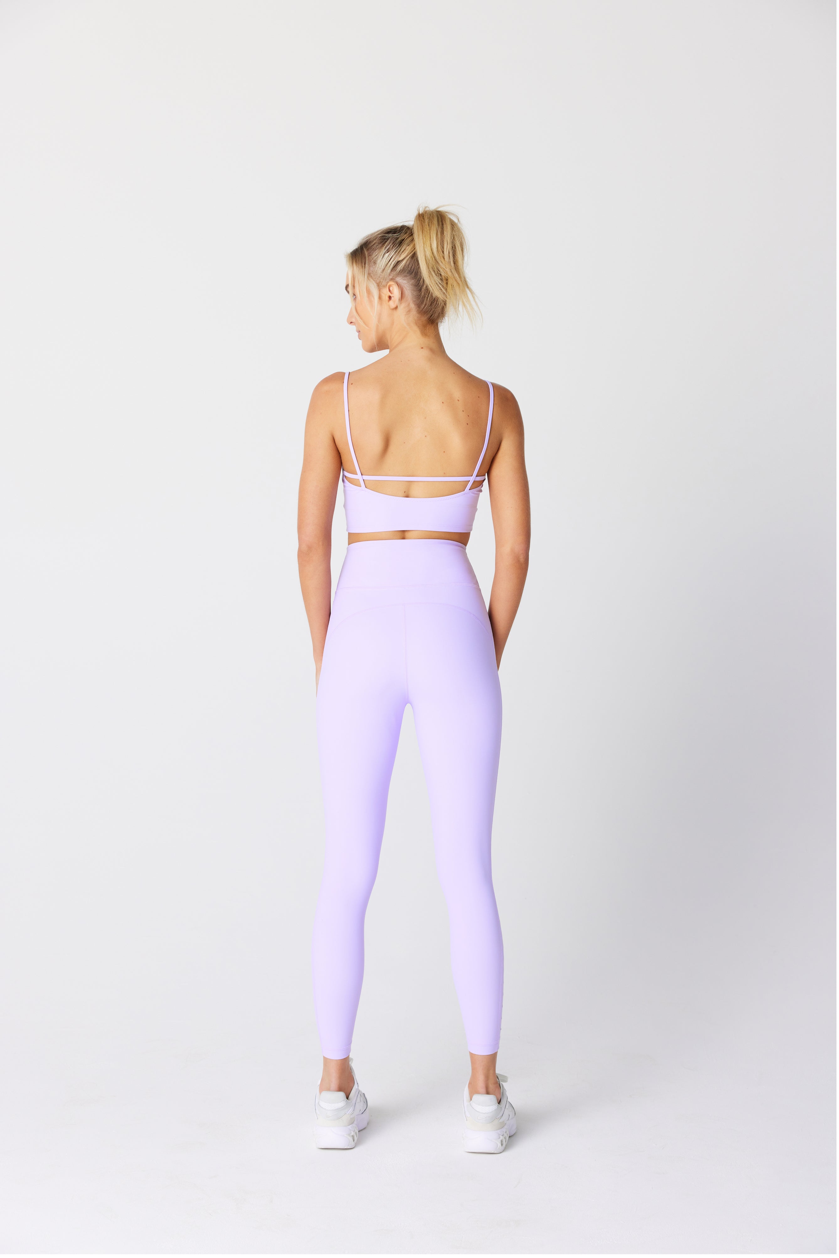 A person with blonde hair in a ponytail wears the pastel purple Avie Top with laser cutouts from Gigi C Sport and matching leggings, complemented by white sneakers. Standing on a plain white background, they face away with hands by their sides.