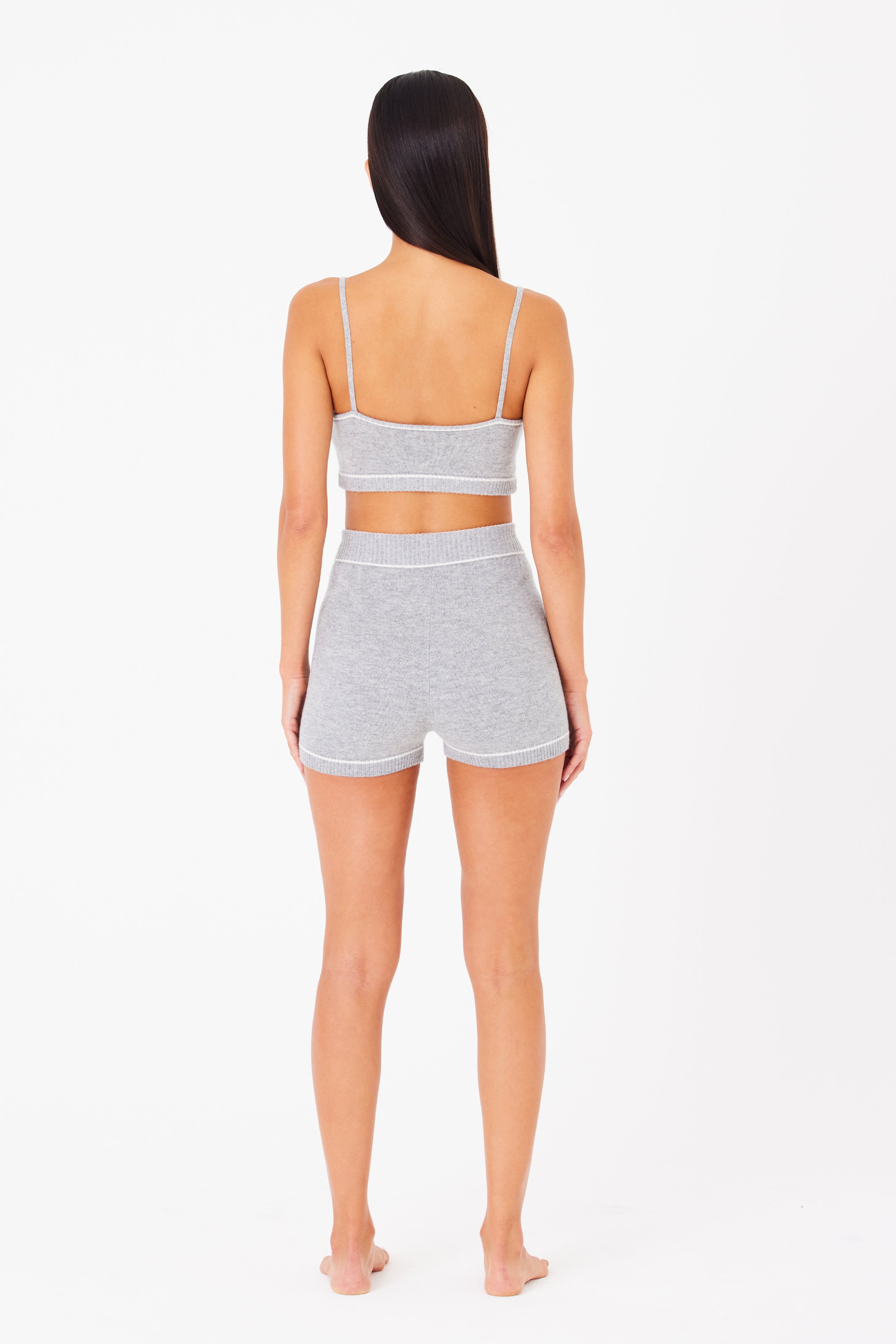 A person with long dark hair models a luxurious, gray loungewear set inspired by the Danica Bralette from Gigi C Lounge. Barefoot and facing away from the camera against a crisp white backdrop, they wear a stylish crop top and high-waisted shorts.