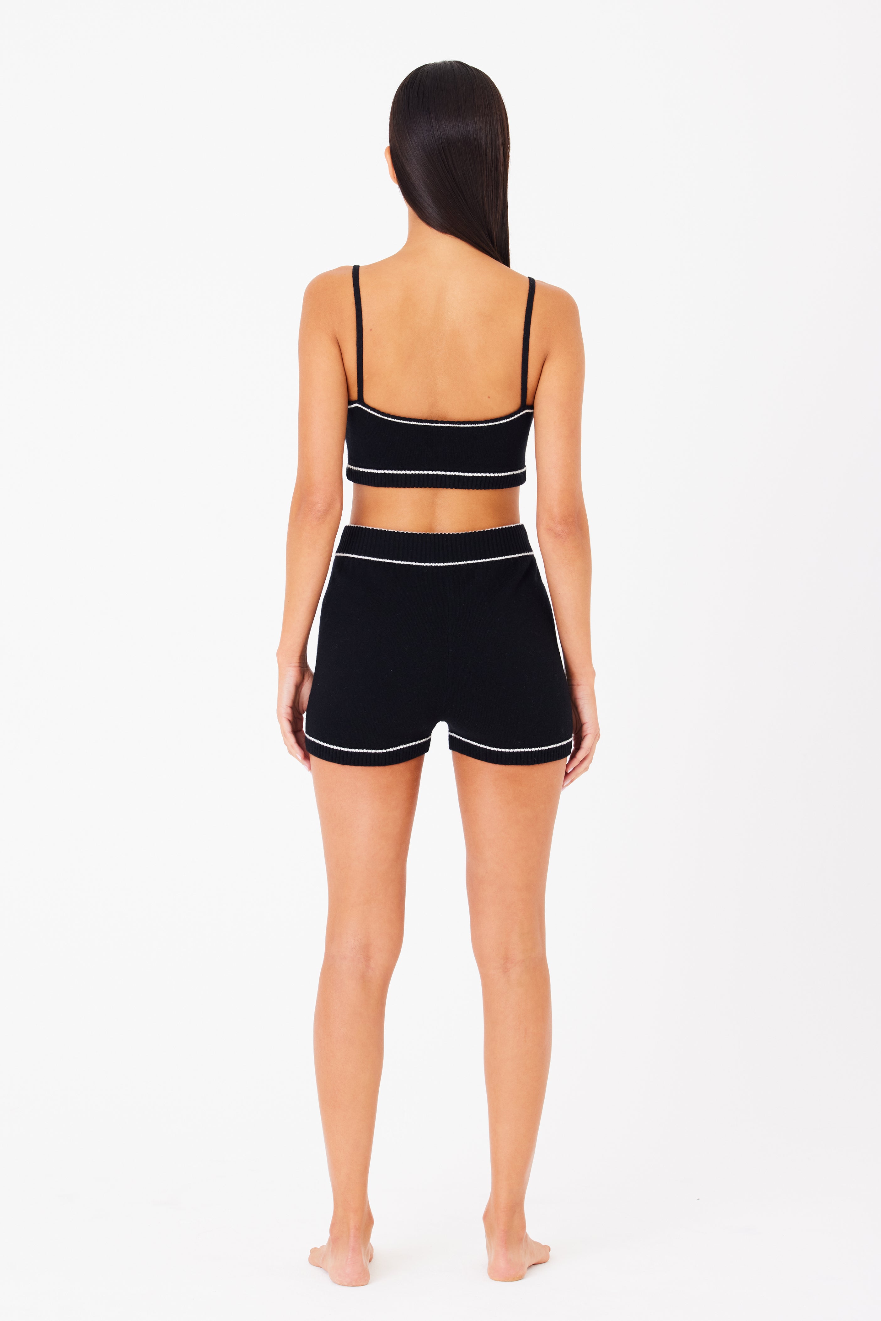 A woman stands barefoot, facing away in a sleek black crop top similar to the Danica Bralette by Gigi C Lounge and shorts featuring white stitching. Her long, straight hair cascades down her back against a plain white background.
