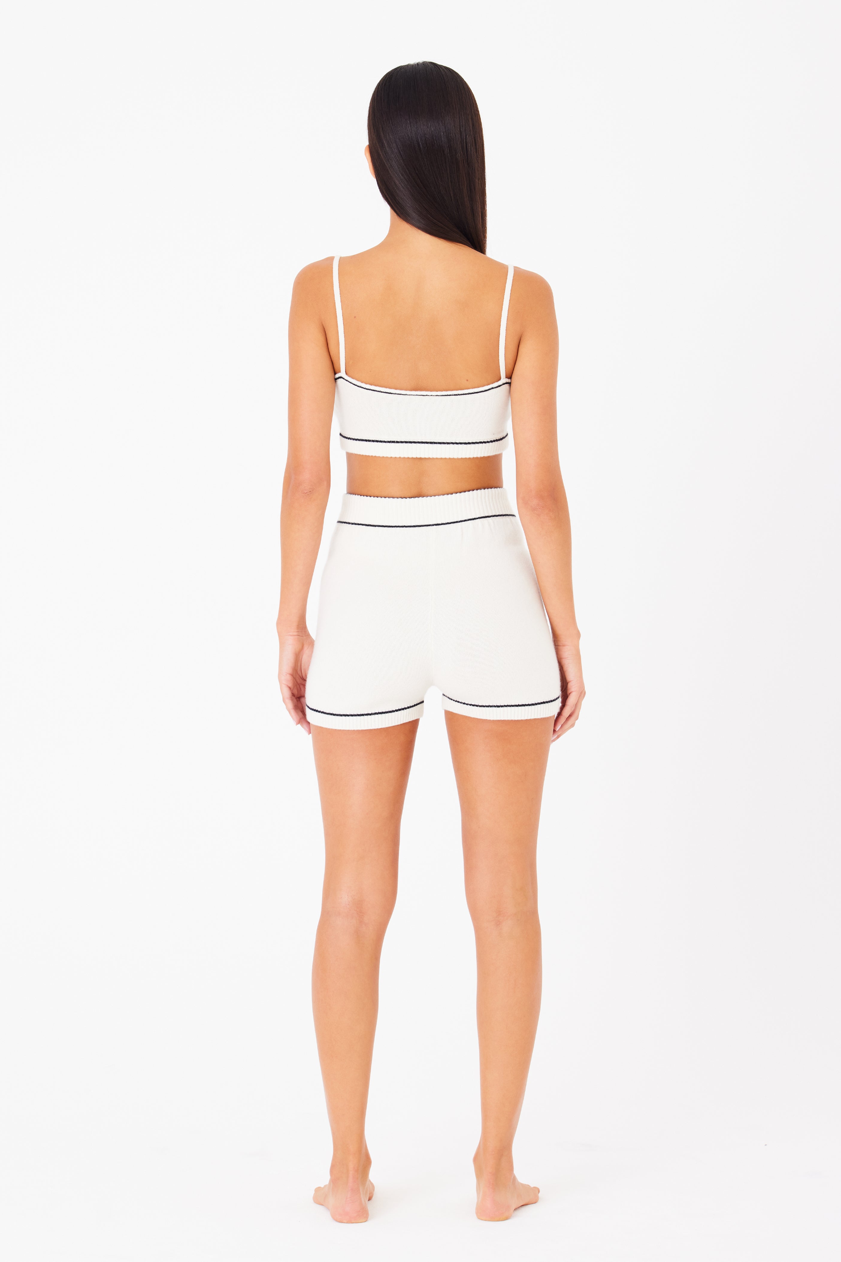 A barefoot individual with long, dark hair stands facing away in a white knit Danica Bralette and high-waisted shorts by Gigi C Lounge. The wool cashmere blend, accented with black trim, offers a luxurious loungewear appeal against the plain white background.